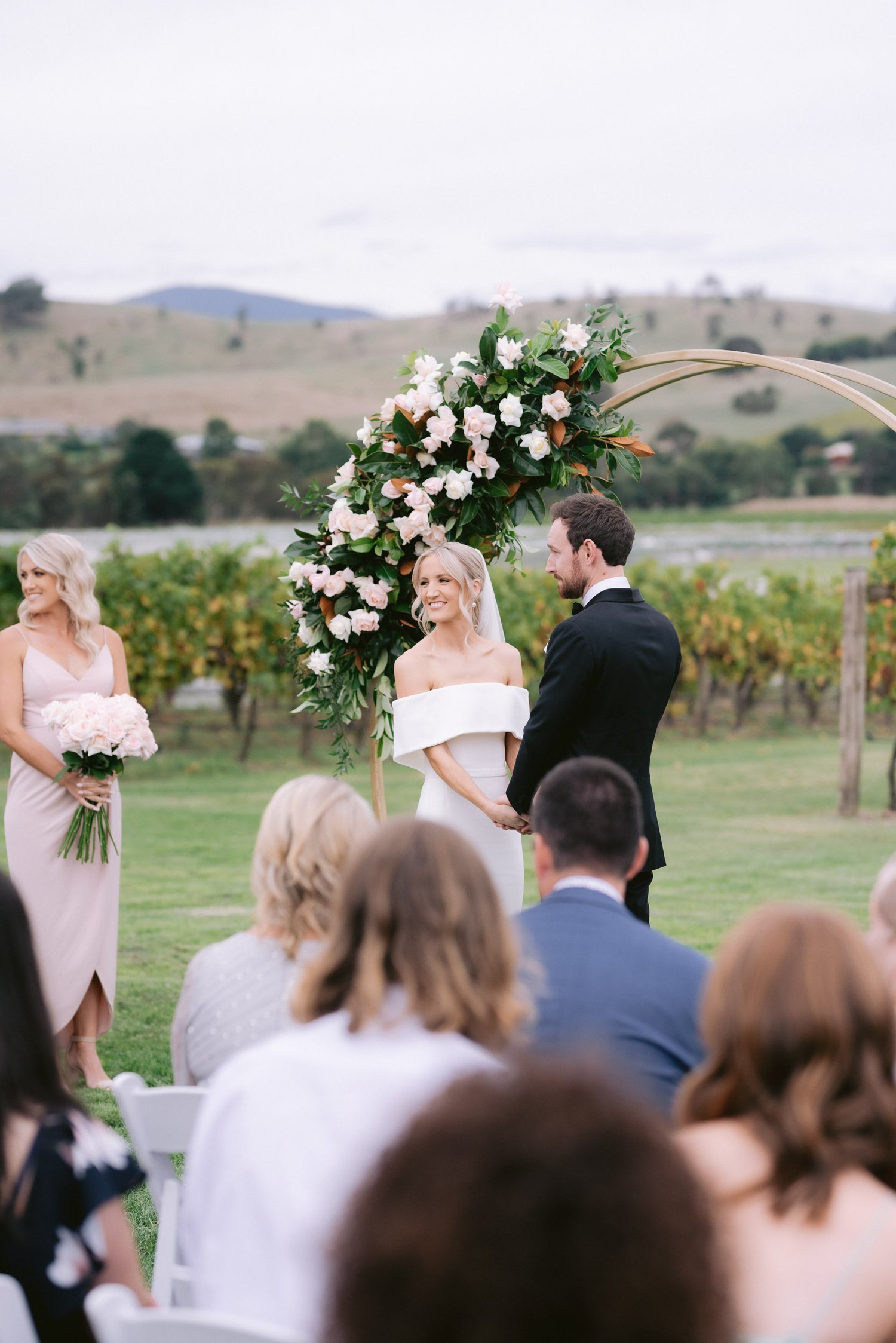 Outdoor wedding ceremony with a bride and groom standing under a floral arch, surrounded by guests seated on white chairs, and a bridesmaid holding flowers on the side.