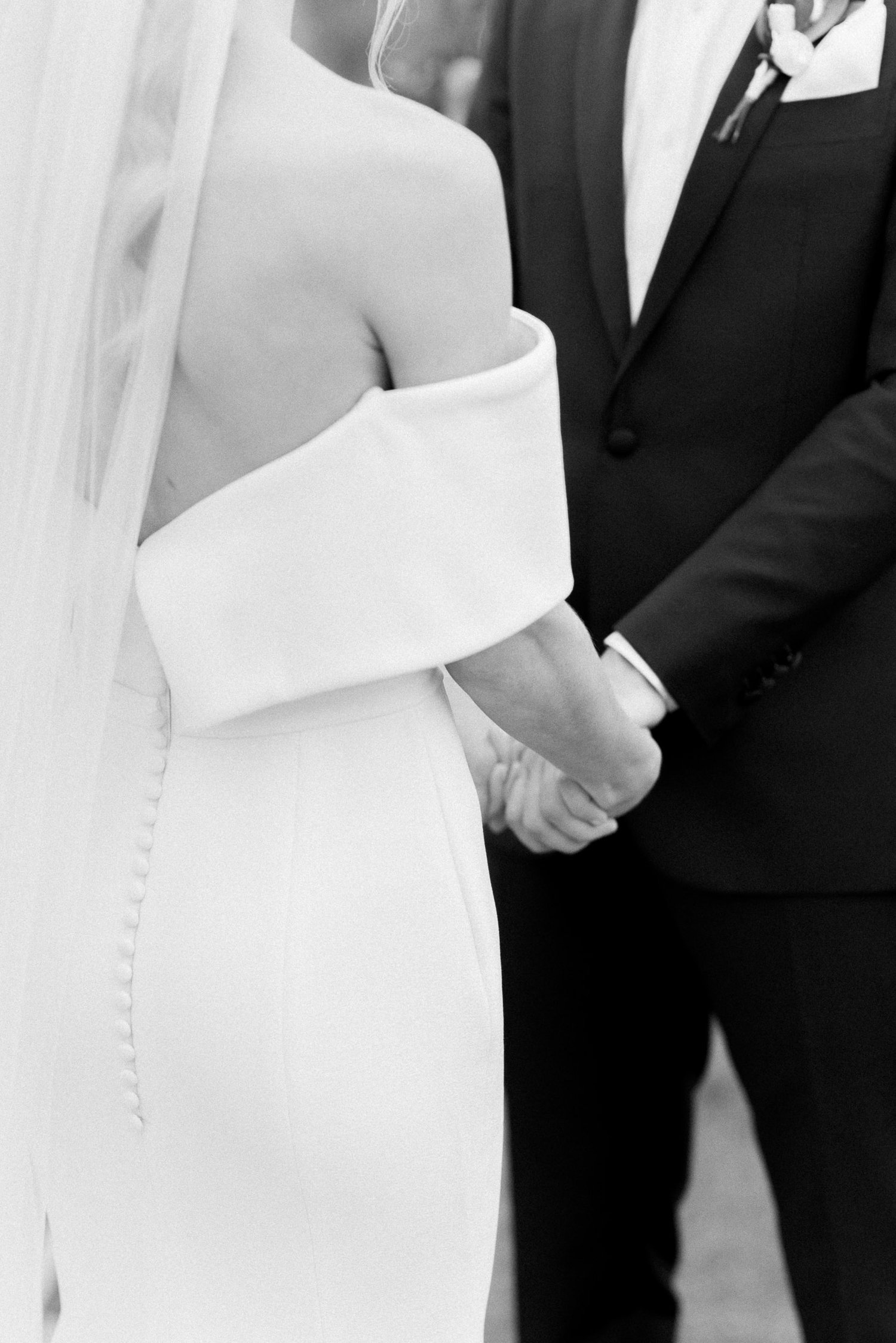Black and white photo of a bride in an off-shoulder wedding dress holding hands with a groom in a suit.