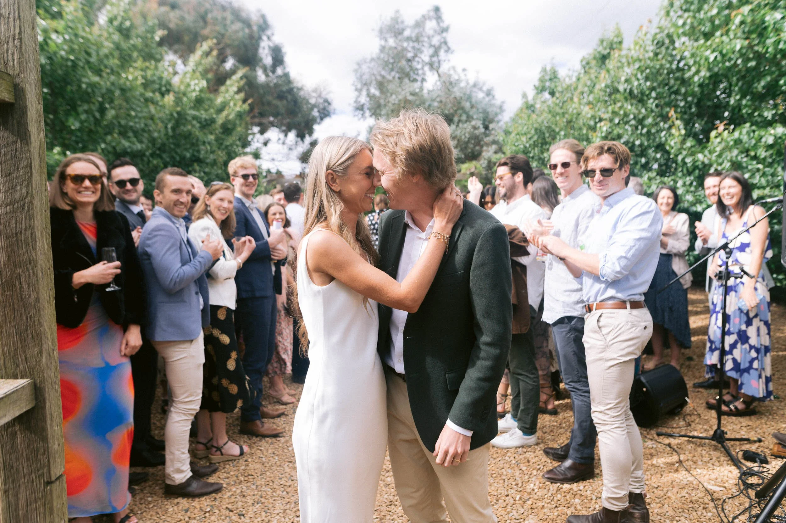 A wedding reception outdoors at Quirindi Stables in Newlyn Victoria with a bride and groom dancing and kissing in the foreground, surrounded by smiling guests clapping and celebrating.