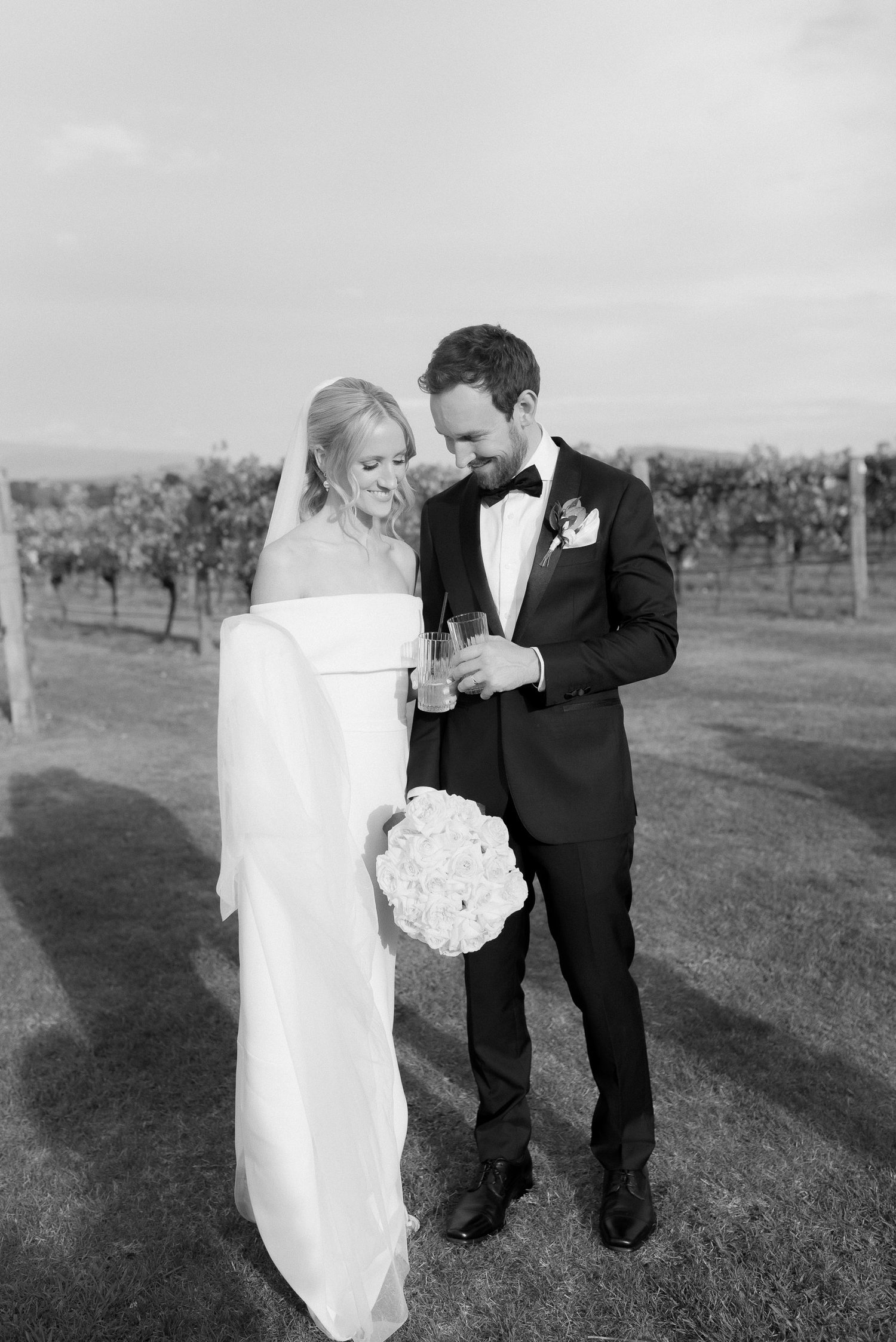 Bride and groom in formal attire holding drinks and a bouquet, standing on grass with vineyard background.