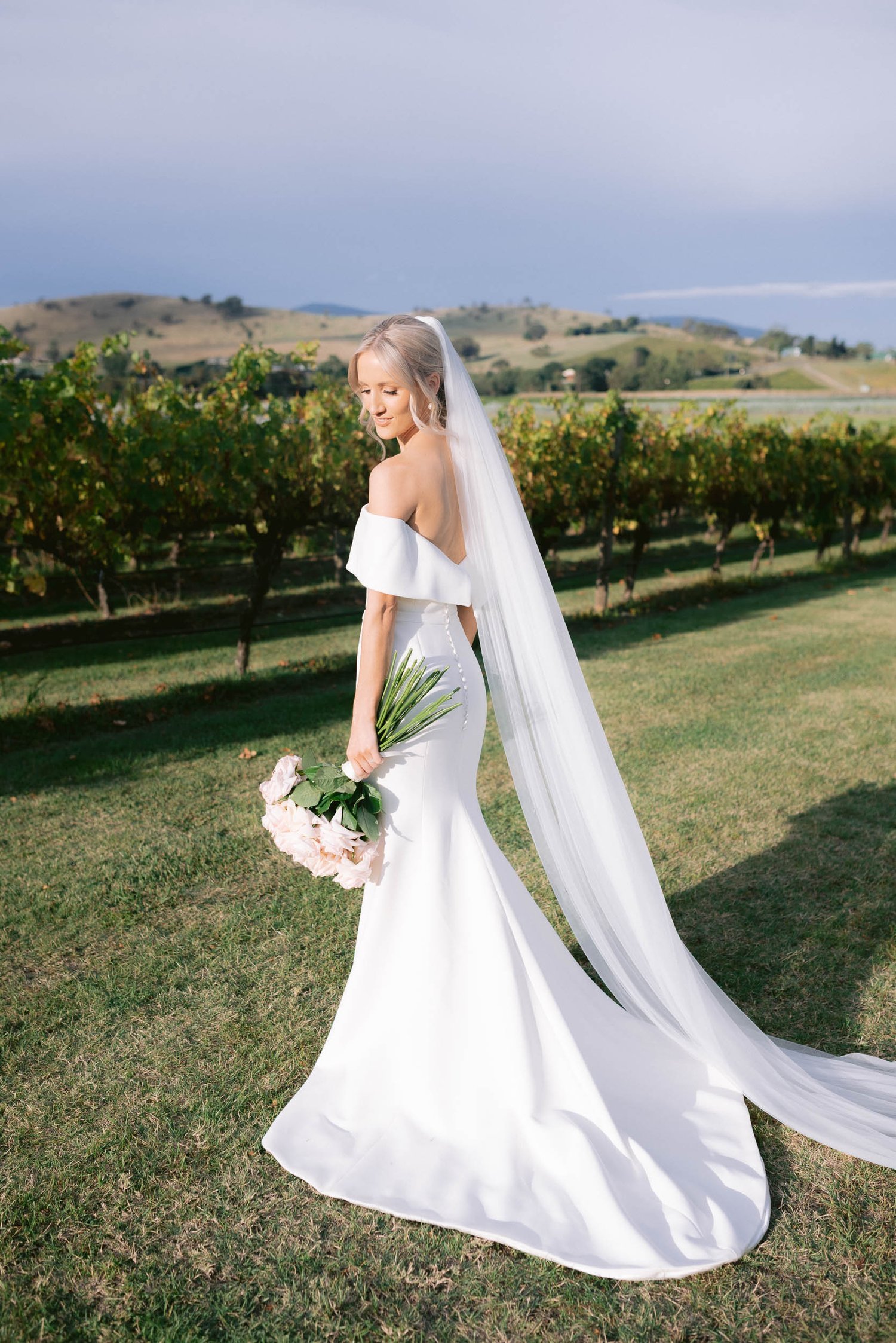 Bride in white dress and veil holding bouquet, standing in vineyard with hills in background.