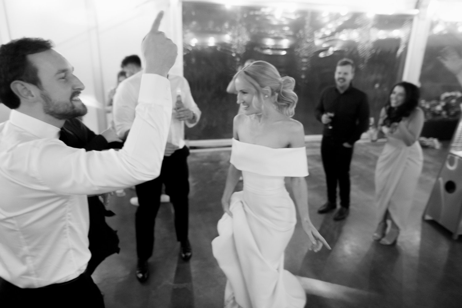 A bride in an off-the-shoulder wedding dress dancing playfully with a man in a white shirt at a wedding reception, with other guests in the background also smiling and enjoying the moment.