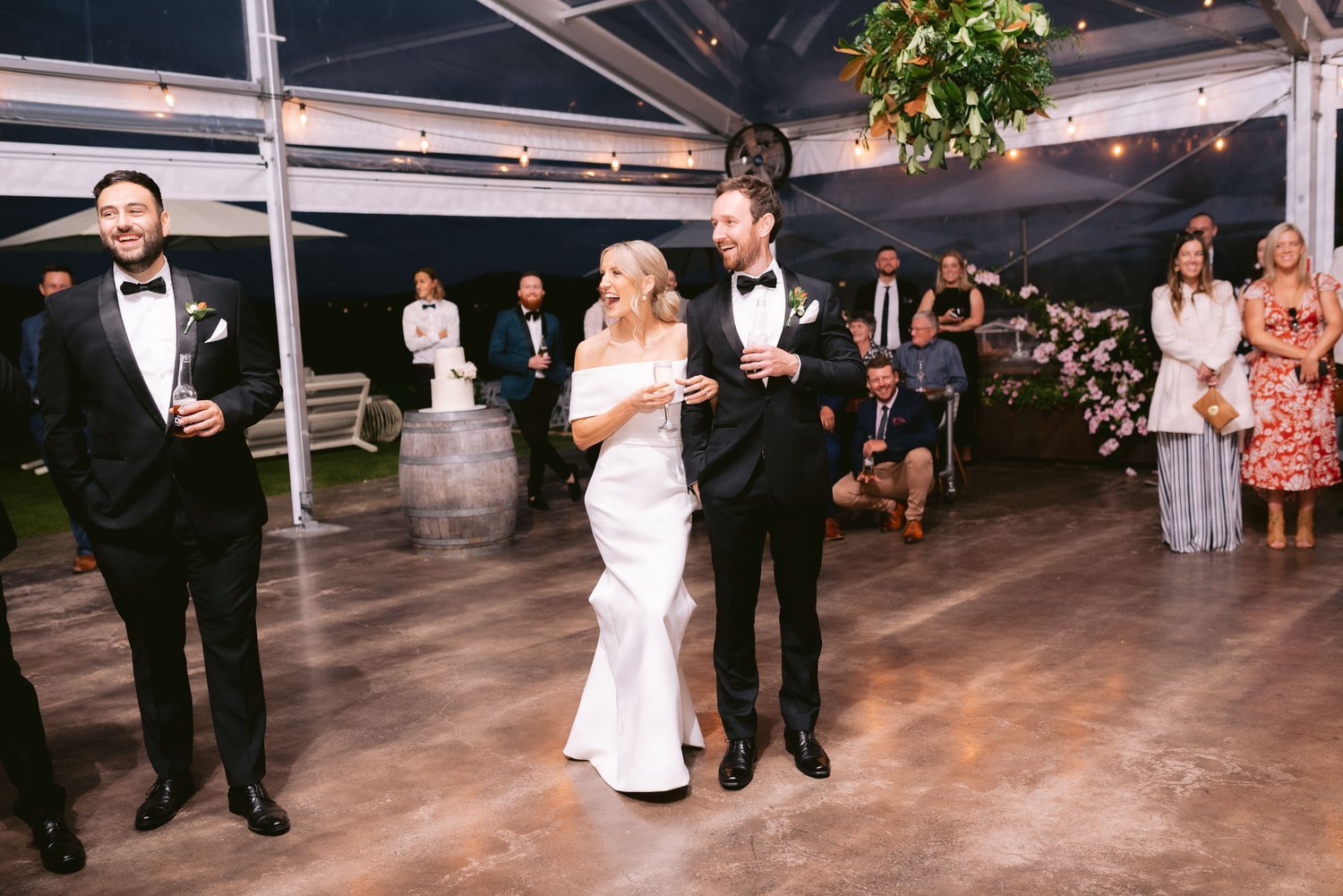 Bride and groom in a white dress and black tuxedo, respectively, smiling at a wedding reception under a canopy with guests watching.