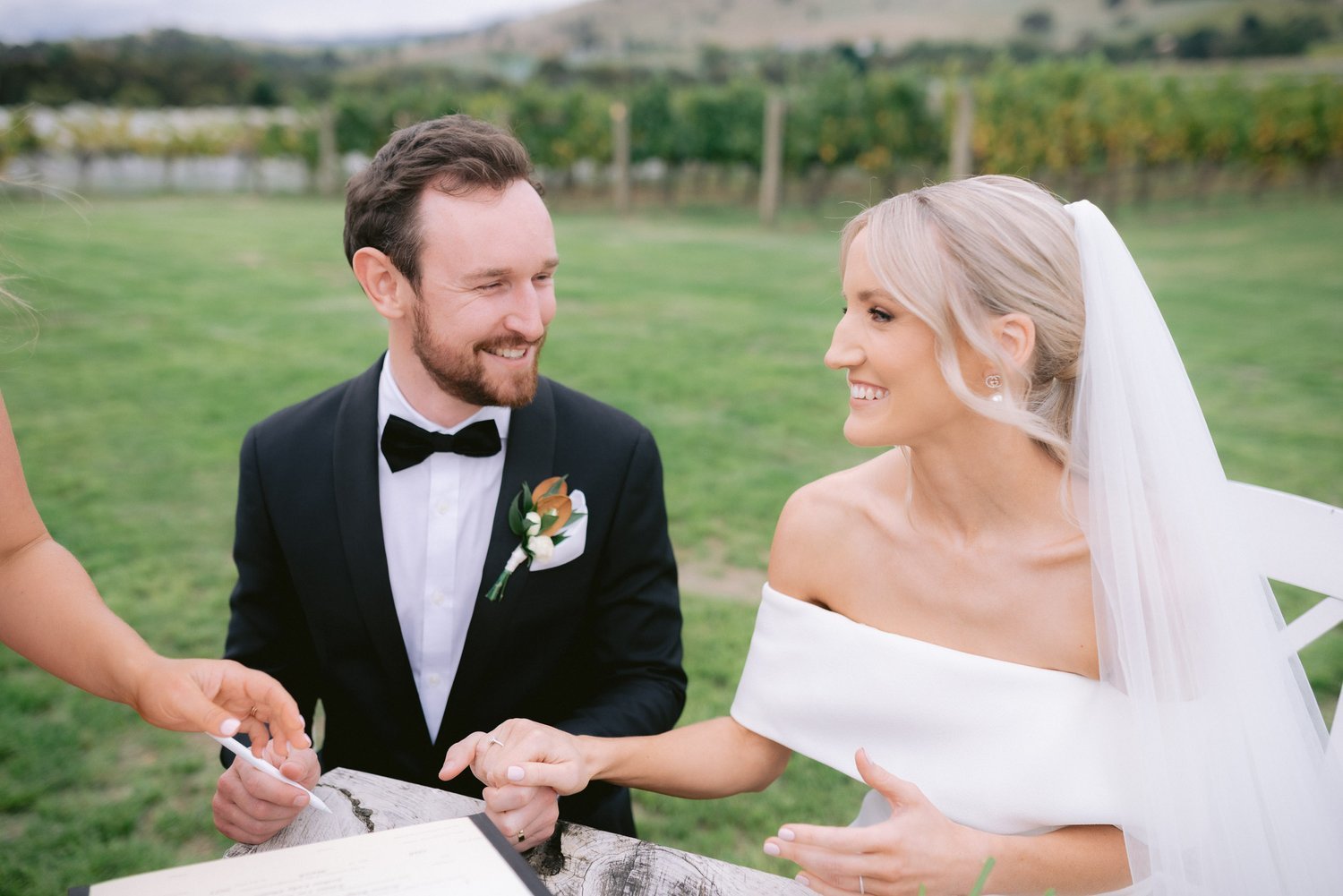 Bride and groom smiling during outdoor wedding ceremony, sitting at a table in a vineyard setting, with a document being signed.
