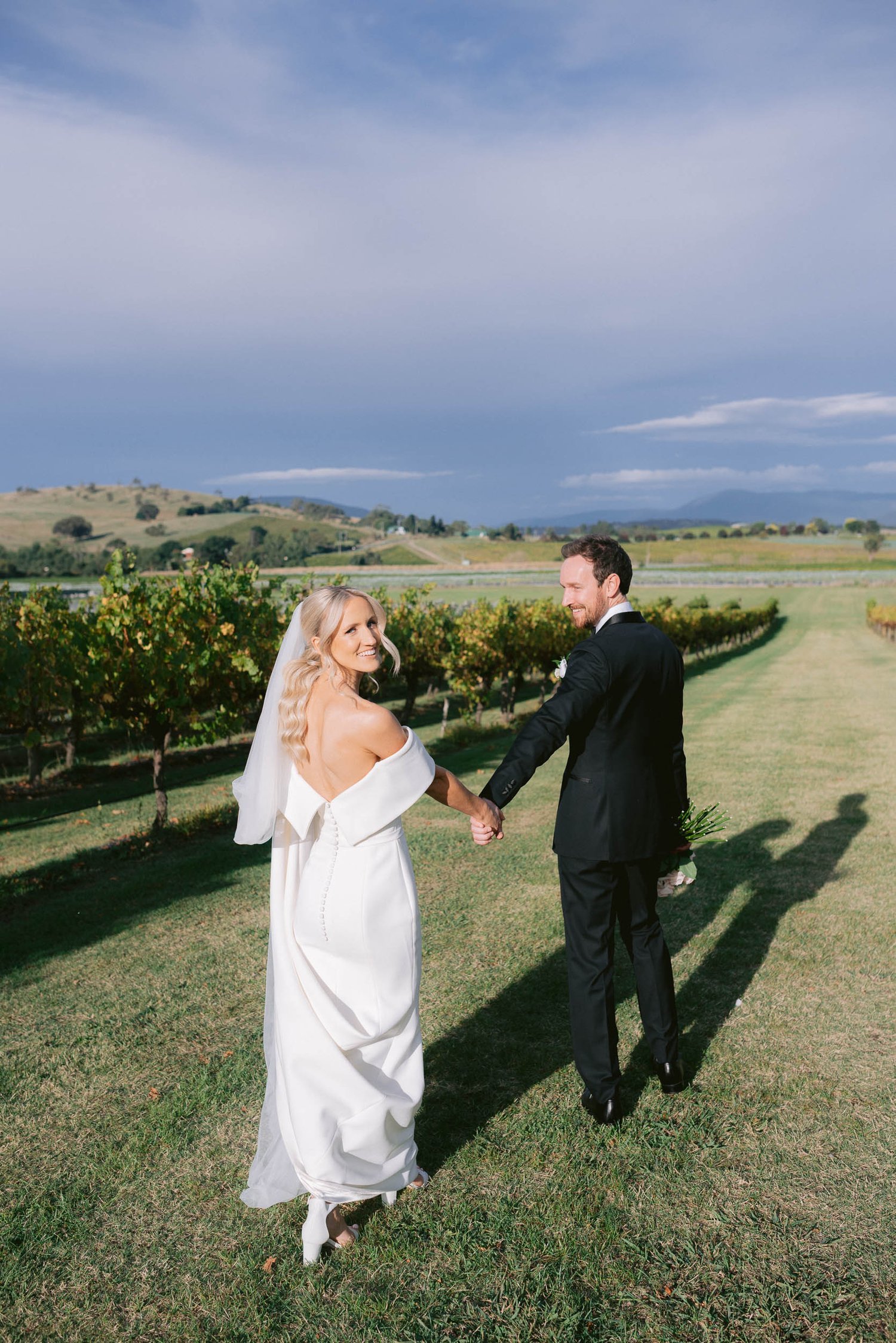 Bride and groom walking hand in hand through a vineyard, with the bride in a white gown and the groom in a black suit.