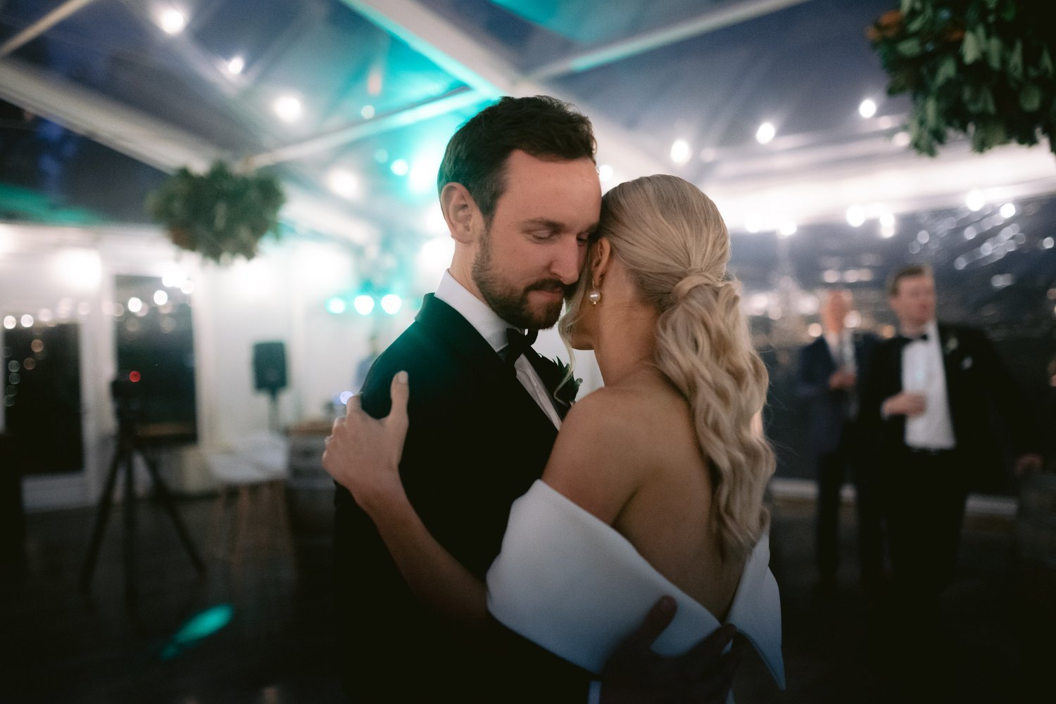Bride and groom embracing on dance floor during wedding reception, with blurred guests and lights in the background.