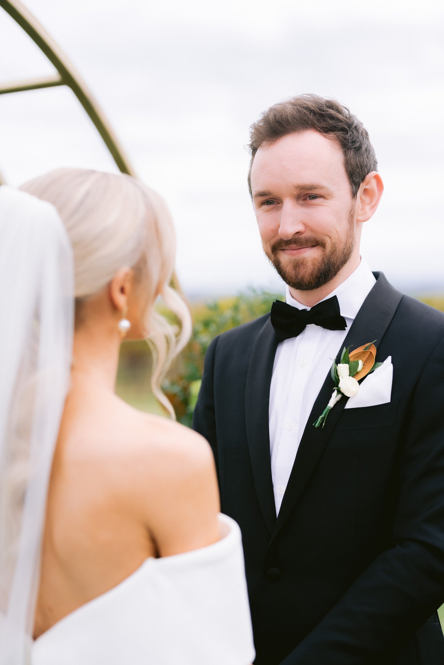 A bride and groom standing face-to-face during their wedding ceremony. The groom is wearing a black tuxedo with a white shirt and a black bow tie, while the bride is in a white off-shoulder gown with a veil. They appear to be outdoors.