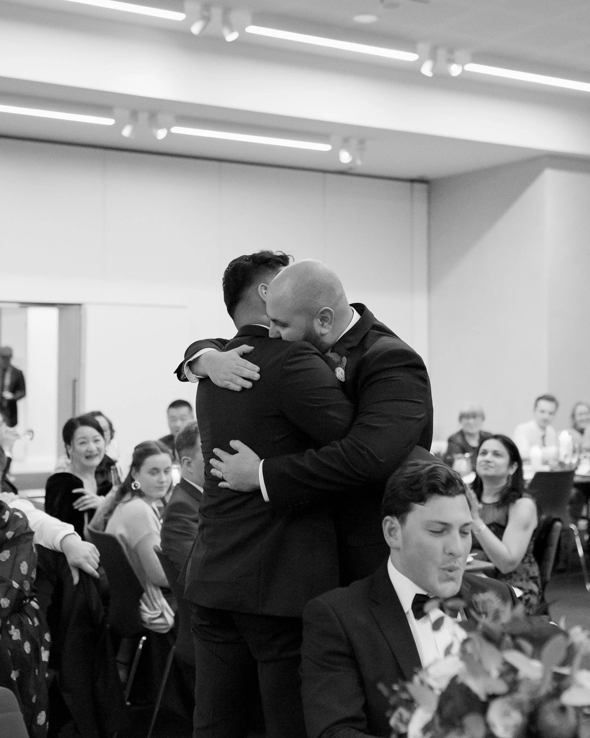 Two men in suits embracing at a formal event with seated guests in the background, black and white.