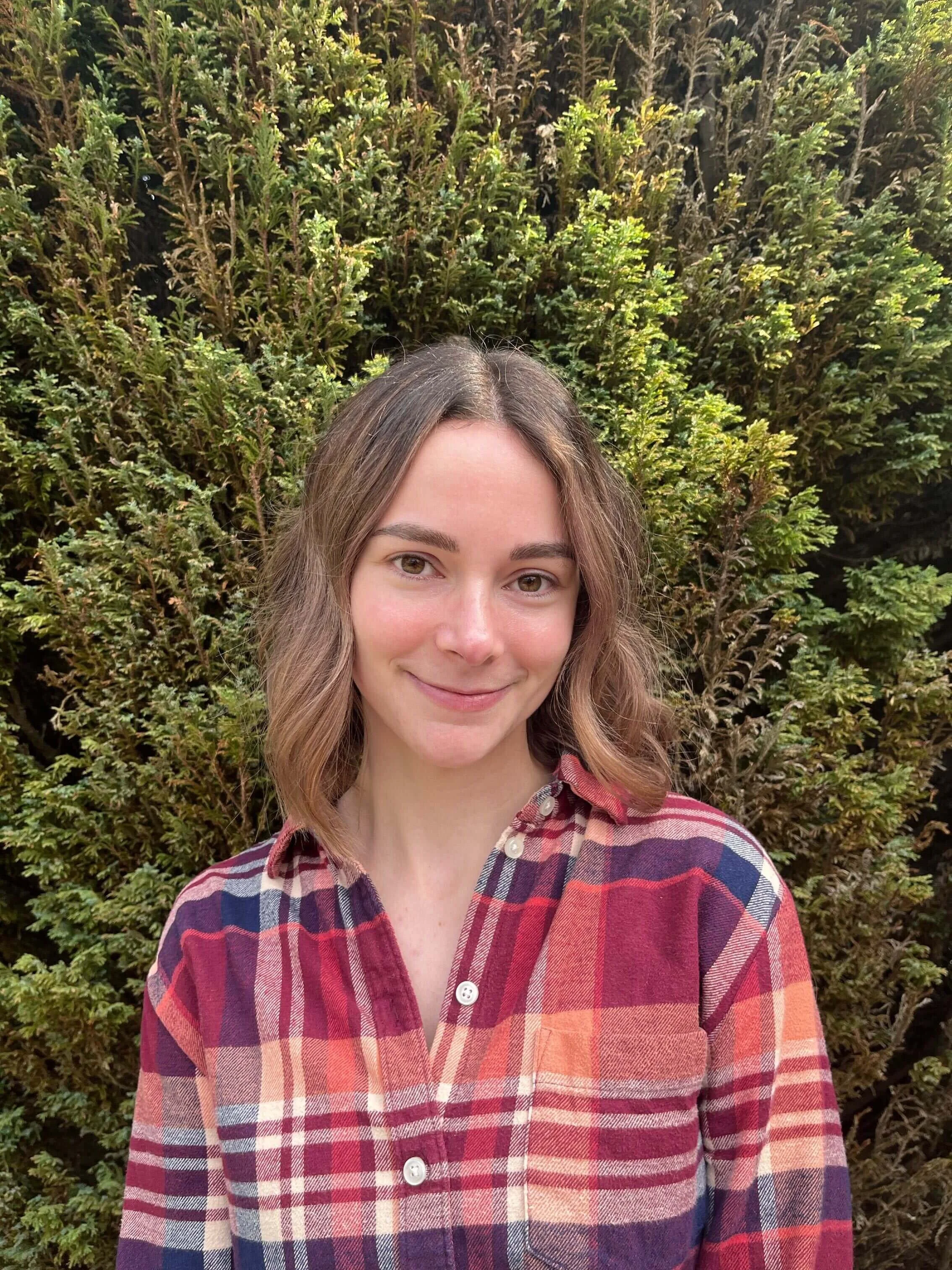 woman with brown hair wearing a pink flannel shirt standing in front of green foliage