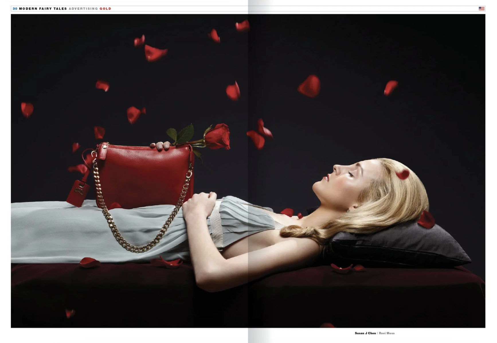A woman with blonde hair lying on her back on a dark surface, eyes closed, with red rose petals falling around her. She holds a red purse with a gold chain and a rose lying on top of it, set against a dark background.