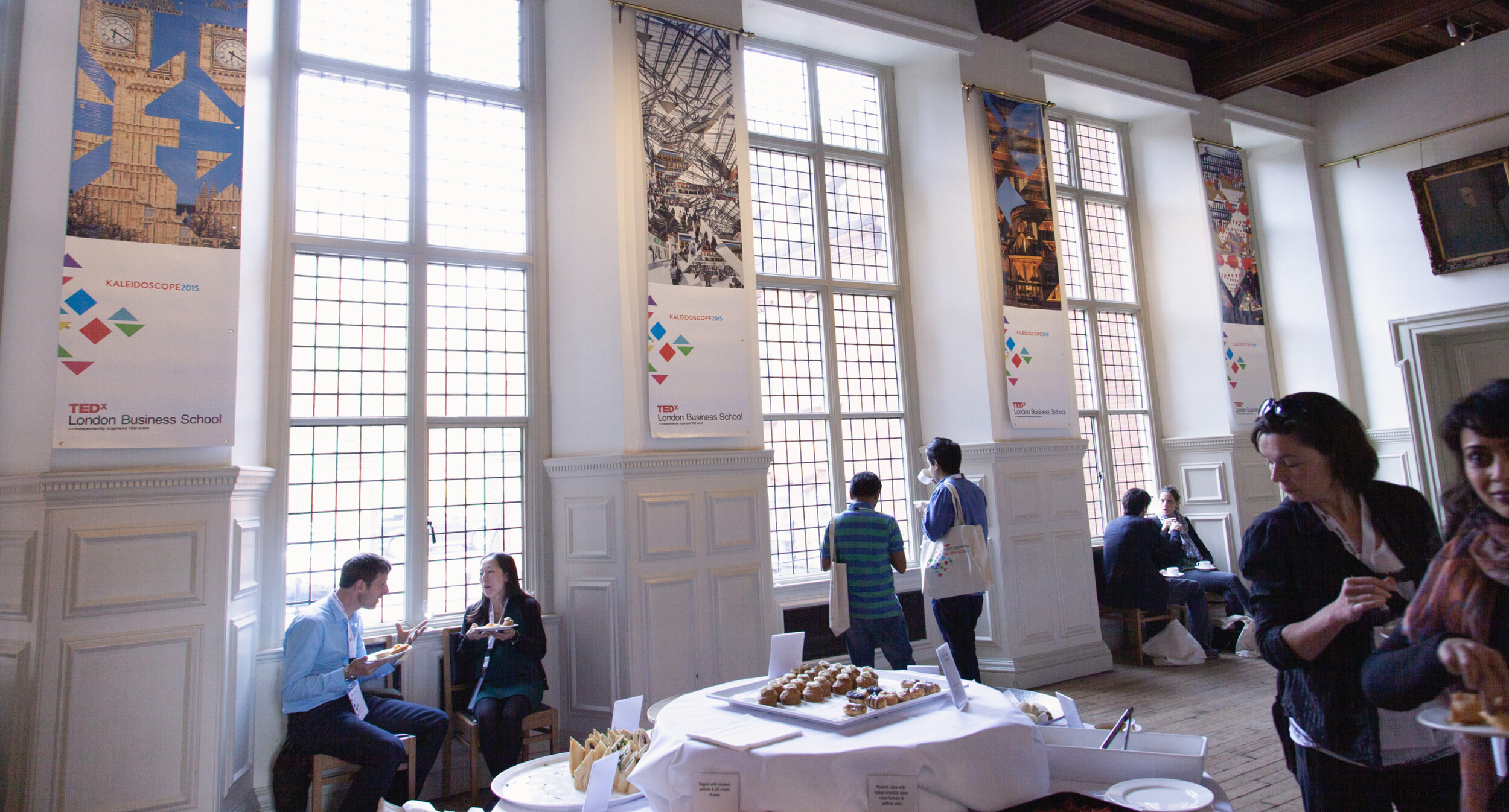 People attending an event at London Business School during a TEDx Kaleidoscope conference, with tall windows, colorful banners, and a table of snacks.