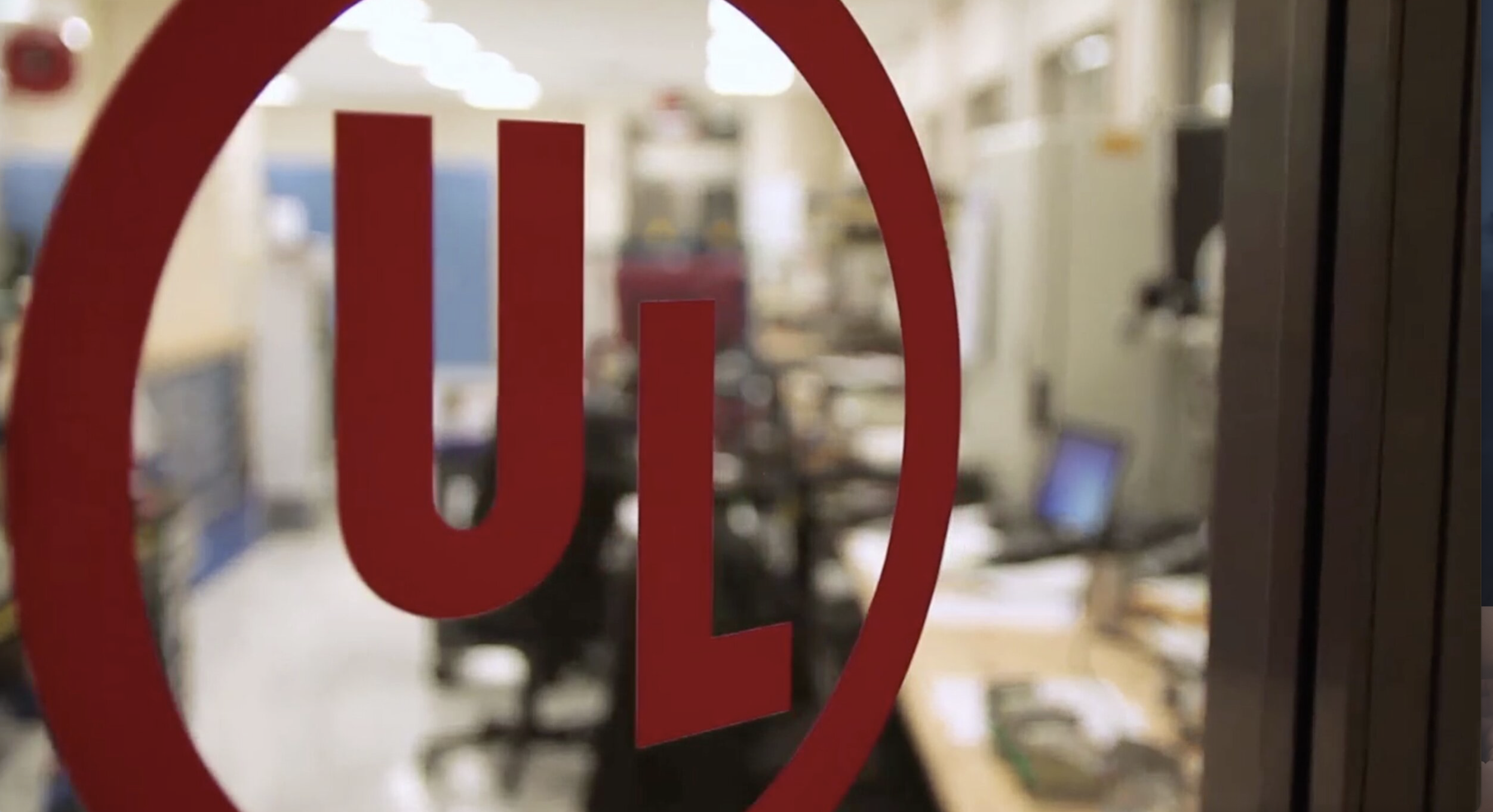Close-up of a red UL Solutions logo on a glass door with a blurred laboratory in the background.