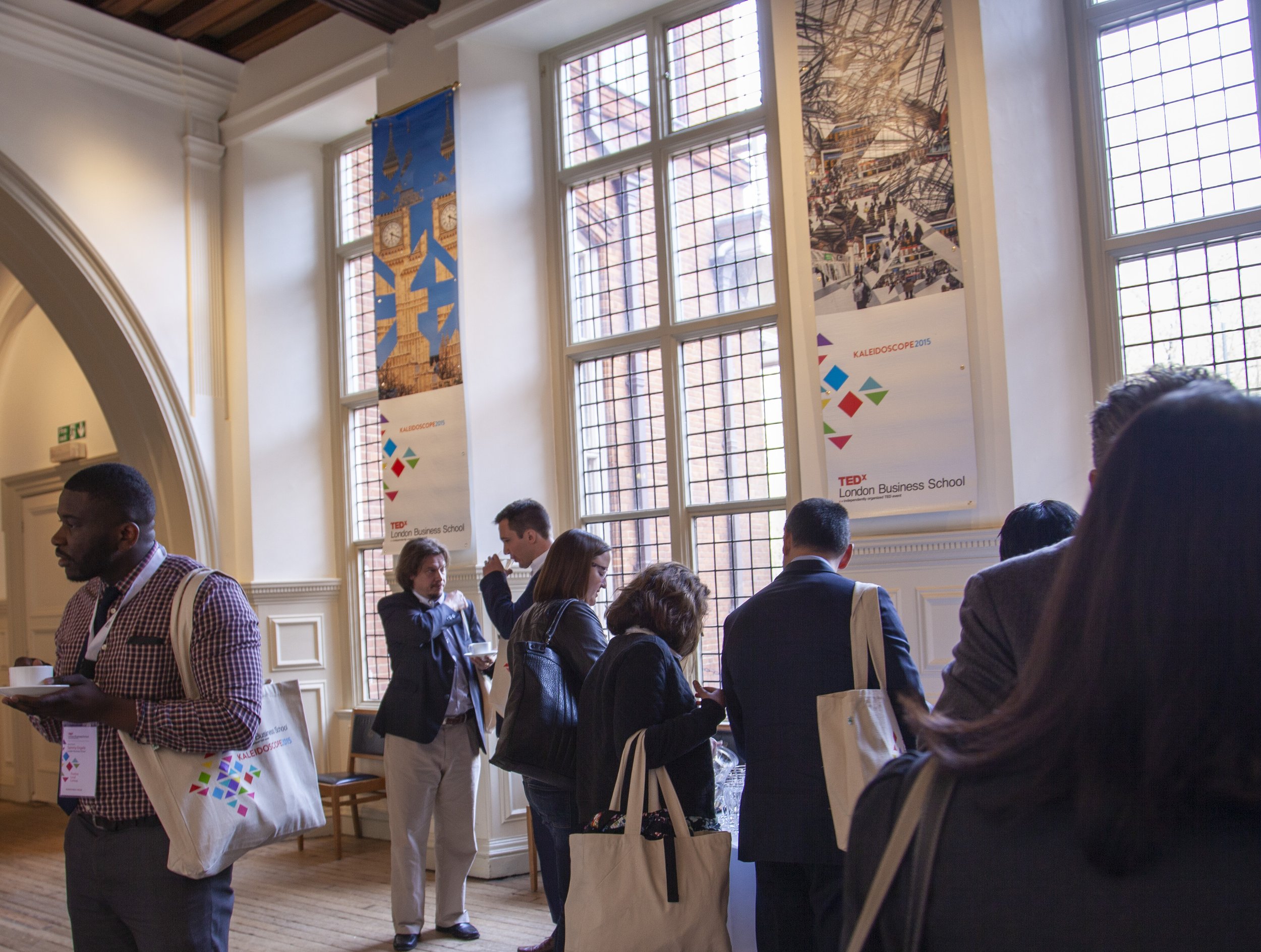 People attending a conference or event, standing and talking in a well-lit room with tall windows and banners displaying event information.