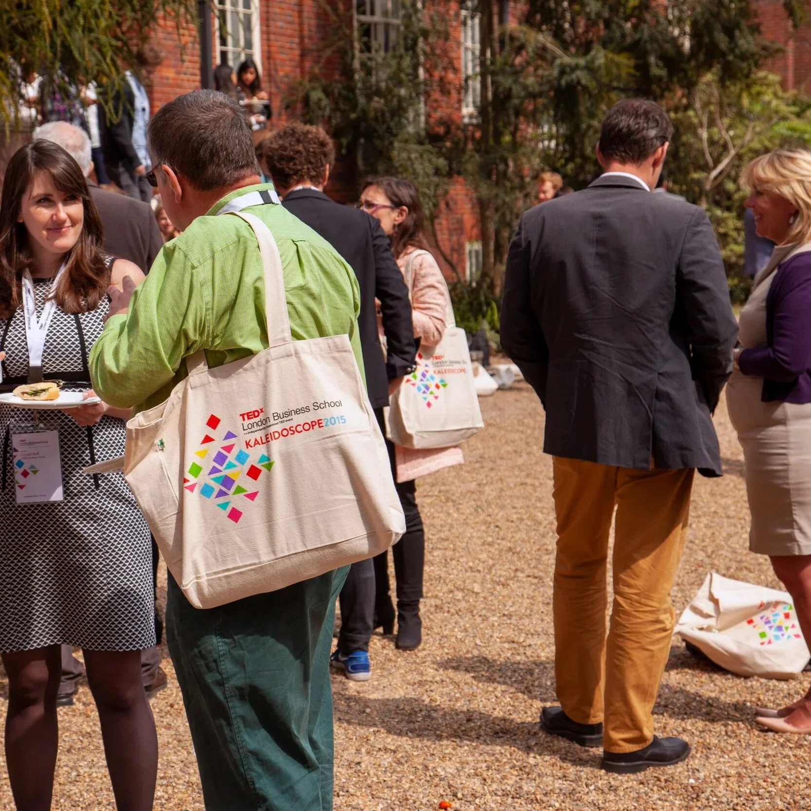 People socializing outdoors at a professional event with several tote bags bearing the TEDx London Business School Kaleidoscope logo. The background features red brick buildings, trees, and a clear sky.