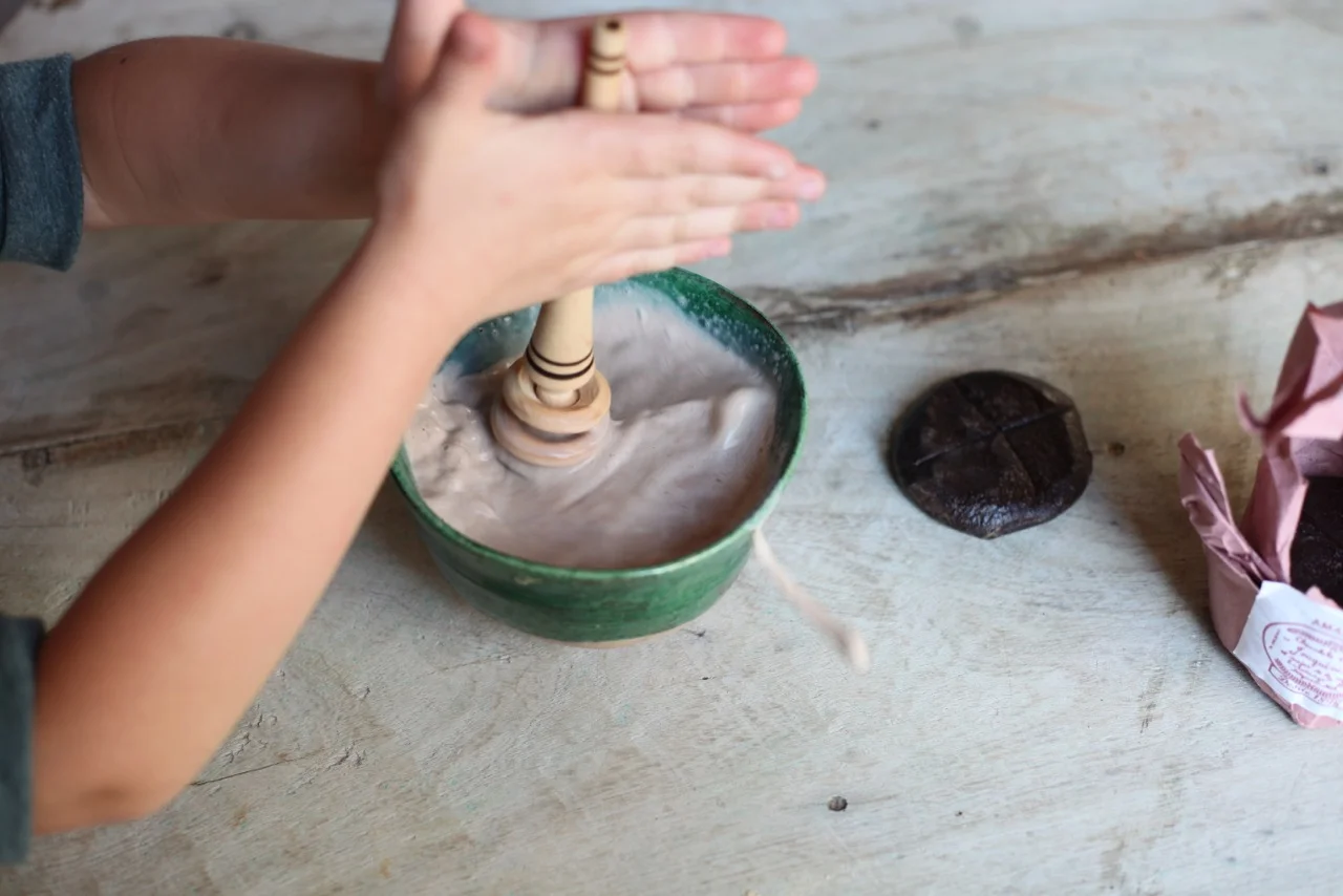 Lola demonstrating the use of the molinillo to incorporate the disk of chocolate into the milk. Note the use of her palms...