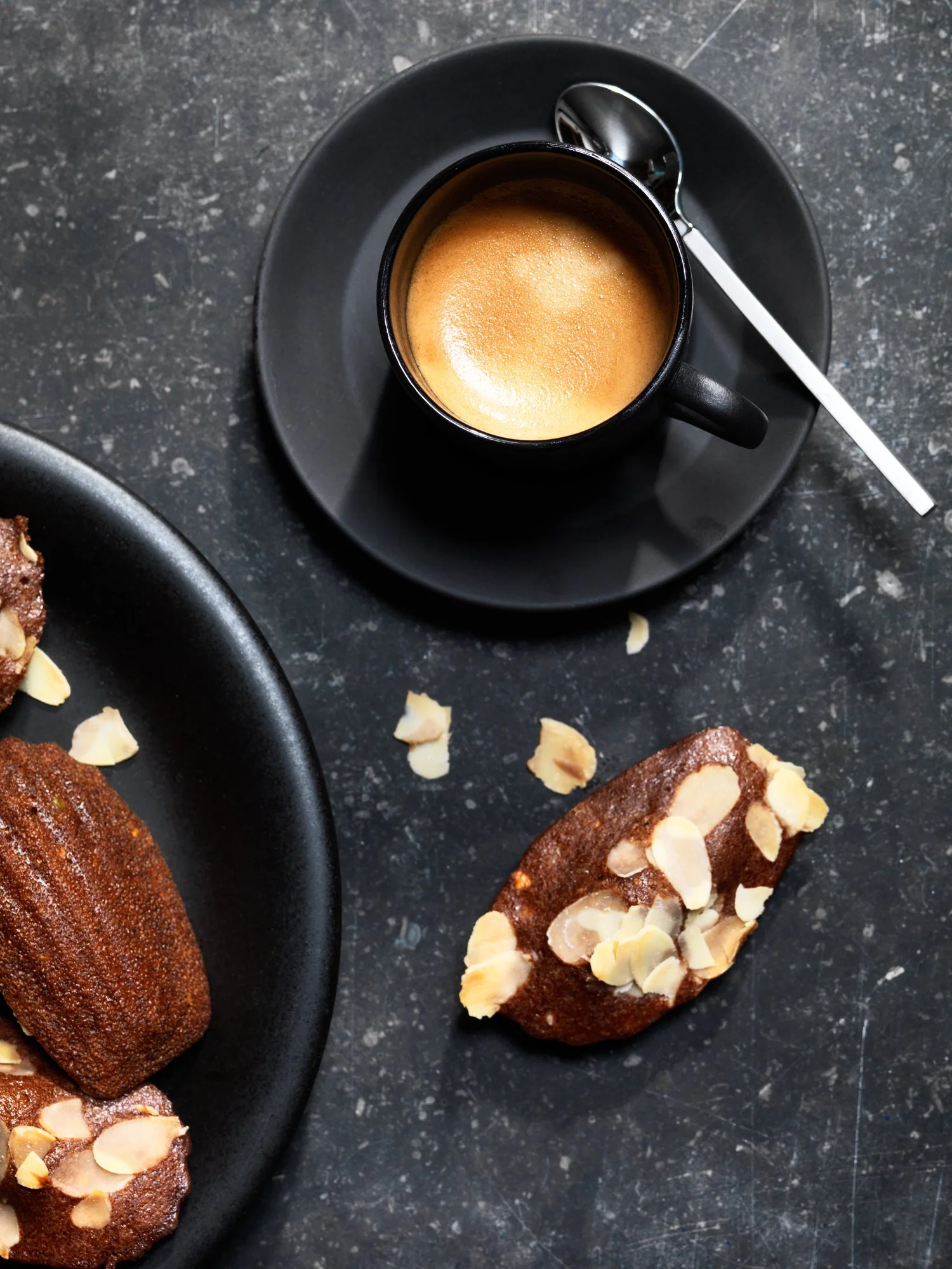 A black cup of espresso on a matching black saucer with a silver spoon, placed on a dark textured surface; partially visible are chocolate almond cakes with sliced almonds on top.