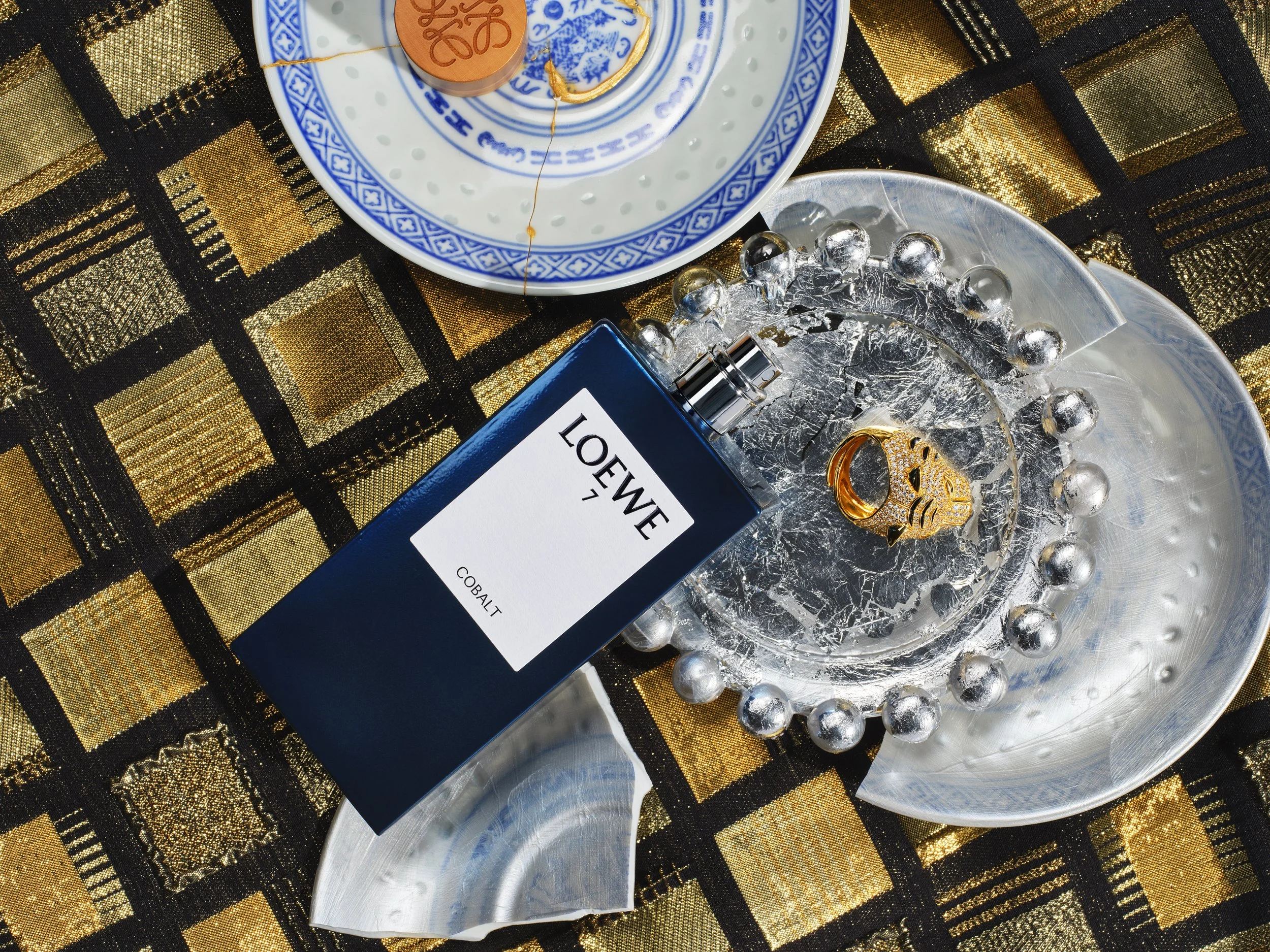 a gold squared table cloth with broken chinese plates on top of that a blue bottle of Loewe perfume and a gold ring