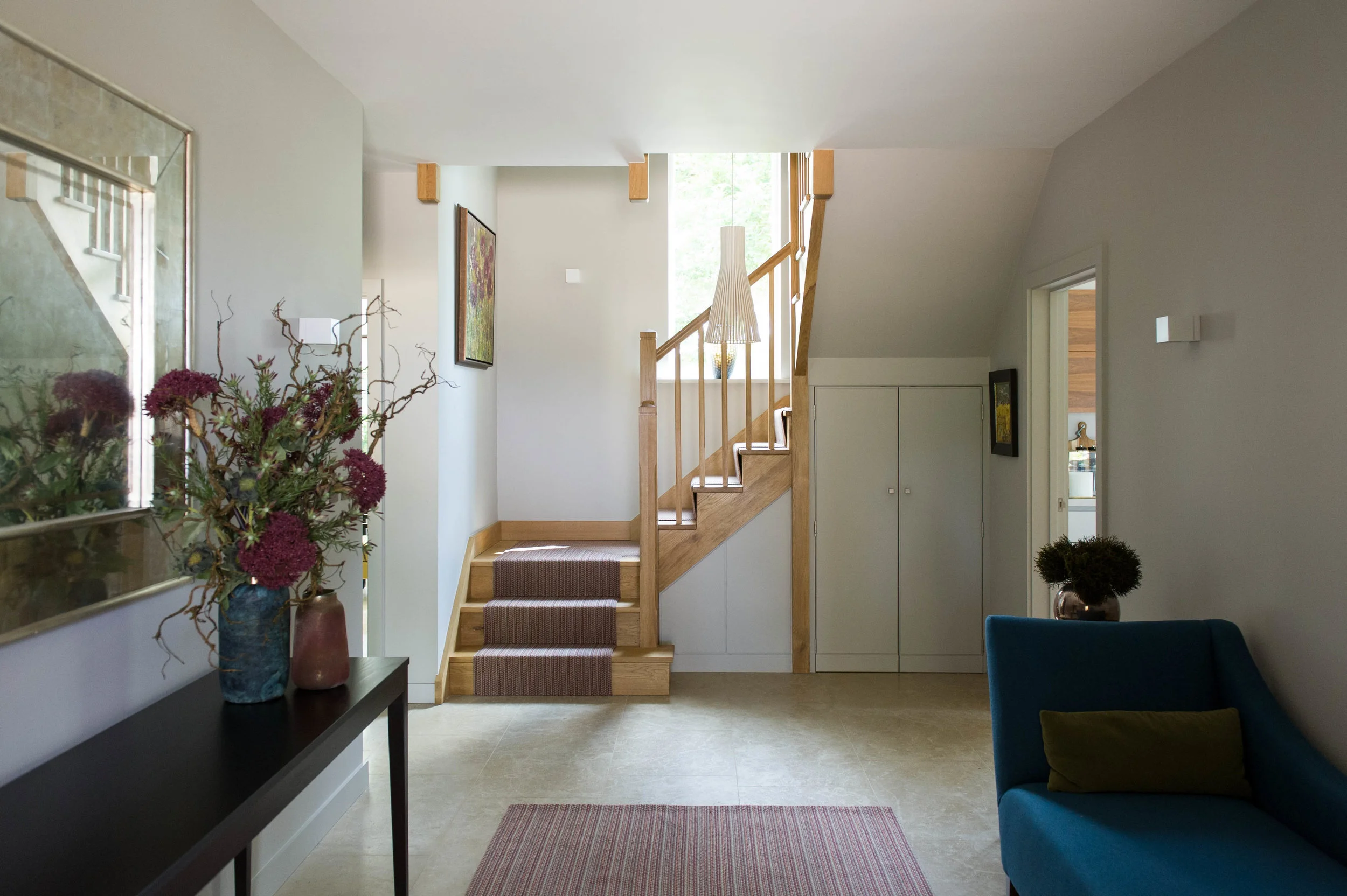 Classic, contemporary hallway with striped rug, chaise longue, oak staircase and antiqued wall mirror above a console table dressed with flowers in a vase. 