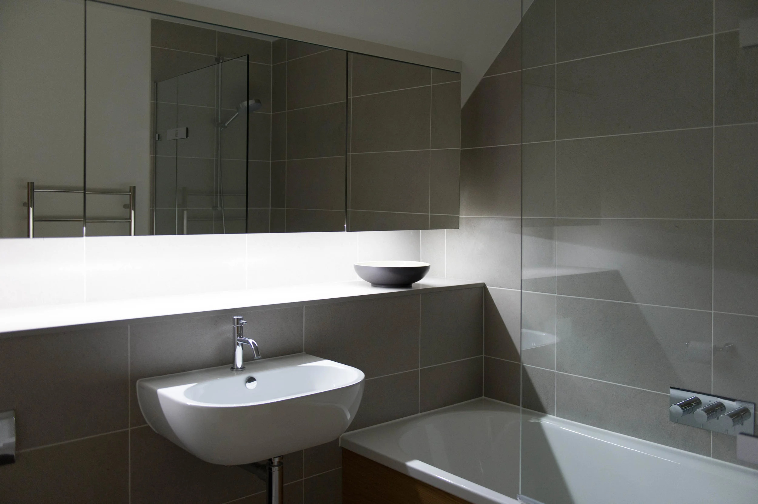 Contemporary bathroom design with white basin and bath, large format grey tiles and bespoke mirrored cabinet.