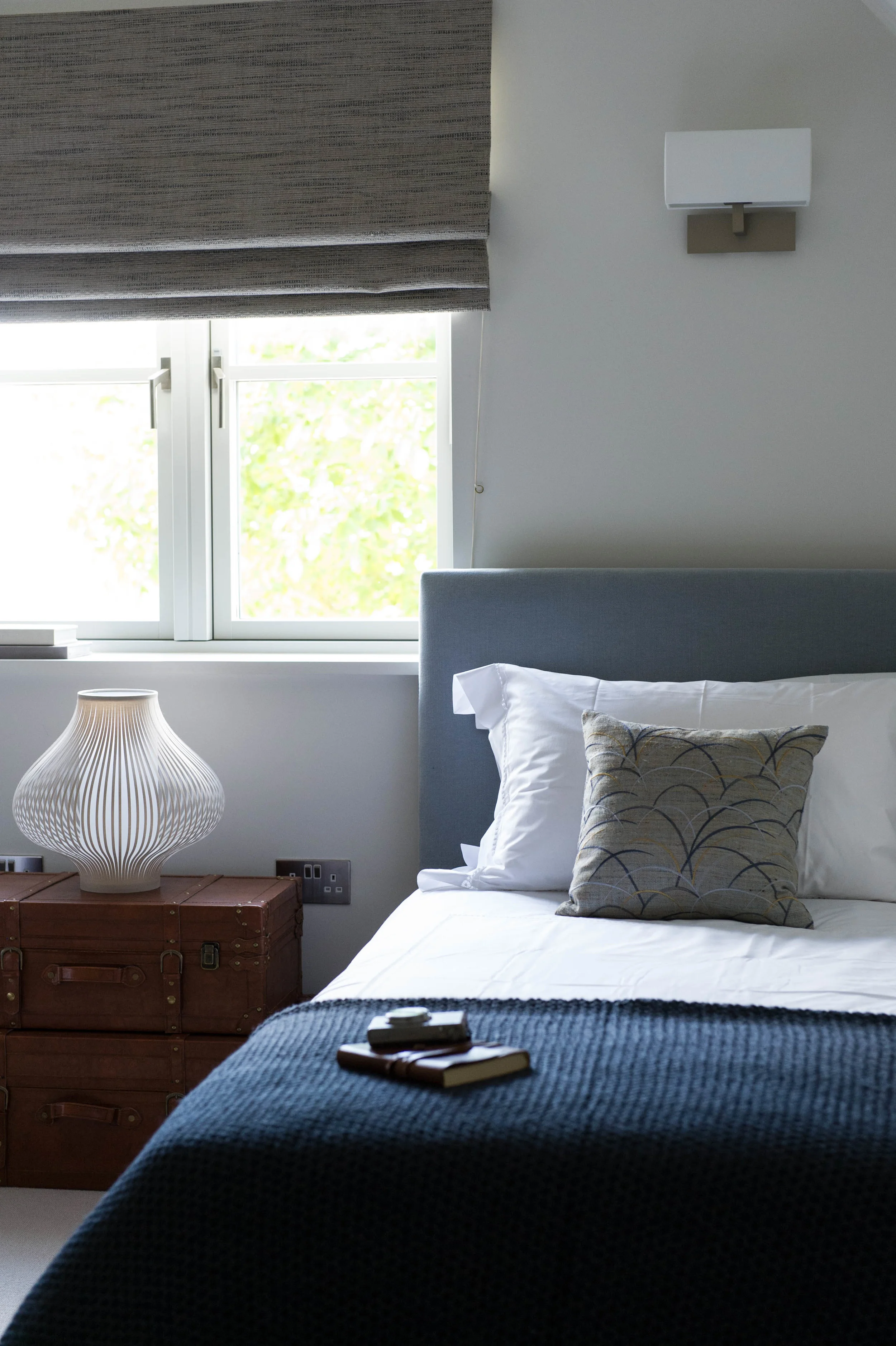 Bedroom with bed made up with navy blue, knitted throw and embroidered cushion alongside pair of leather trunks with table lamp on top.