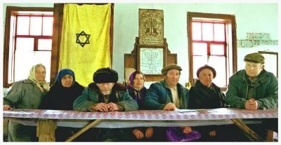 "It has the kind of walls that keep in the cold of winter and the heat of summer," says Bershad Jewish community President Yephim Chaim Vigodner (far right) breathing a good-natured sigh as he surveys his 200 year-old synagogue. The roof and floor sag. A pair of old tefillin (leather phylacteries) grows mold in the rotting podium. Rusty matzah-making machines and decaying volumes of Talmud litter the women's balcony. Yet, miraculously and mysteriously, the synagogue exists, surviving the Cossacks, pogroms, Nazis and Communists. During World War II, the Jewish population of the ghetto surrounding the synagogue swelled to nearly 30,000, as the Rumanians, Moldovans and Ukranians shipped their Jews to this shtetl (Jewish village) seven hours from Kiev. Today, of Bershad's14,000 inhabitants, fewer than 100 are Jews. On Shabbat, 20 of those healthy enough among the remaining community kiss the centuries-old mezuzah on the synagogue doorframe and gather on the creaky benches before the original ark. The problem is that after decades of Communist repressions, no one in the community can read Hebrew or recollect Jewish prayers. The last rabbi left in 1956. One frigid Friday night, an out-of-town visitor occasions a larger crowd than usual. Huddling with them, he teaches ancient Jewish melodies without words – for some, it is the first service they have ever seen. The community and synagogue seem to shed layers of dust and for an instant, their voices rise to the sky. Ironically, it may have been that very same dust which enabled them to hide these many generations. As Yephim says, "Perhaps we were humble enough to be spared."