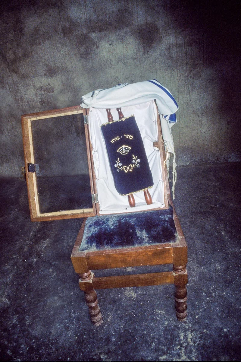The House of Israel community’s miniature Torah is housed in Tefereth Israel synagogue in a glass and wooden case, which rests on the seat of a chair and is typically covered with a tallit. The Torah, carried to the community by Washington, DC-area resident Daniel Baiden, is removed from the ark every Shabbat morning. Though it is tiny, the community loves this Torah.
