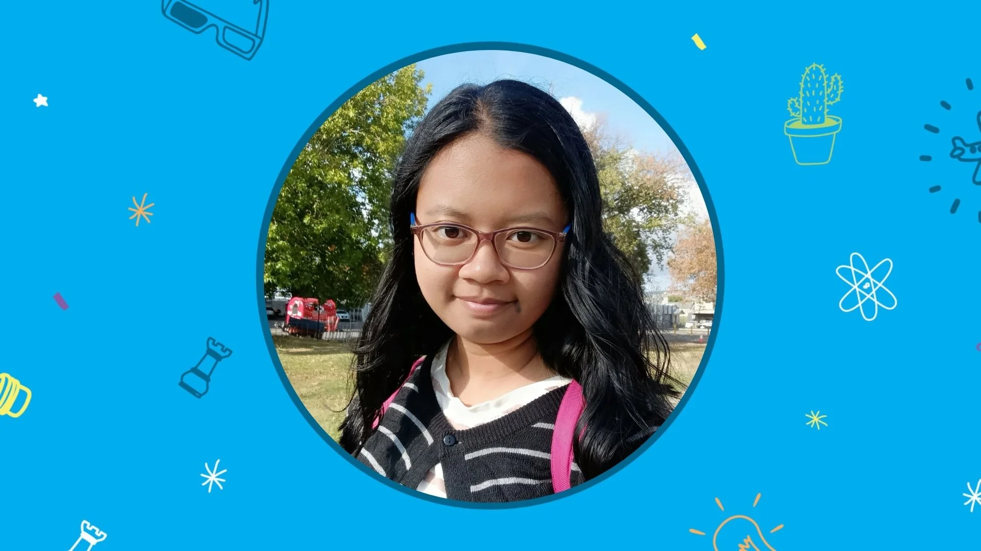 A photo of a young woman with long black hair and glasses. She is smiling into the camera. There are trees in the background hair and glasses