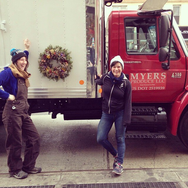Two people posing for a Christmas photo with a wreath on the side of a box truck. 