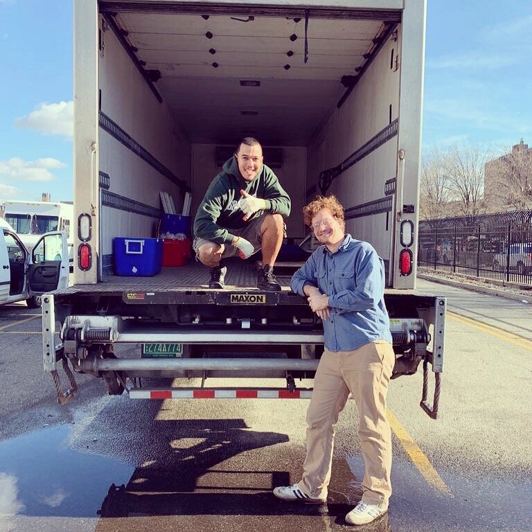 Two people in the back of a box truck  with the back open. They are posing while leaning on the back of the truck. 