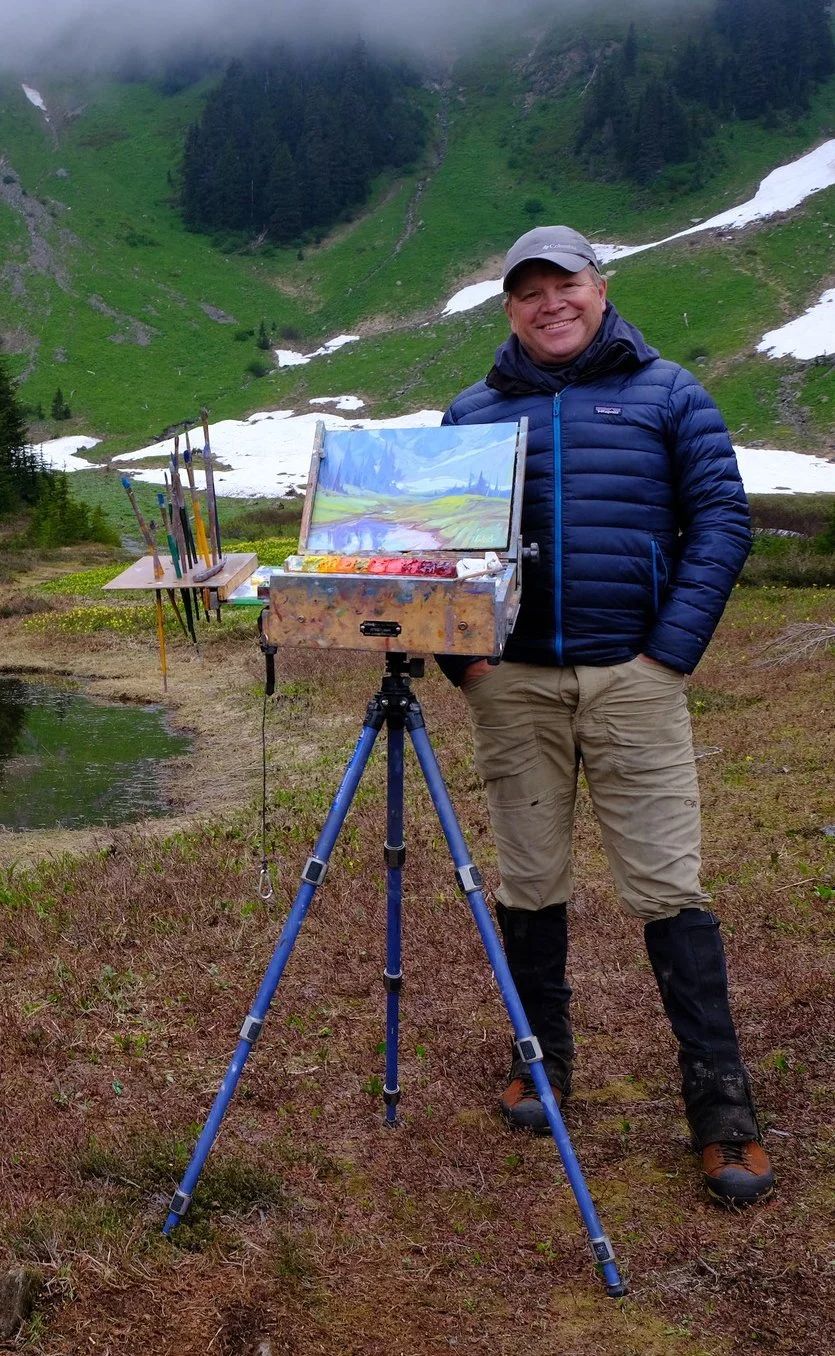 A man standing outdoors in a mountainous area with patches of snow, green hills, and trees. He is smiling, wearing a gray cap and a blue puffer jacket, and standing next to an easel with a painted landscape of a river, green fields, and blue sky.