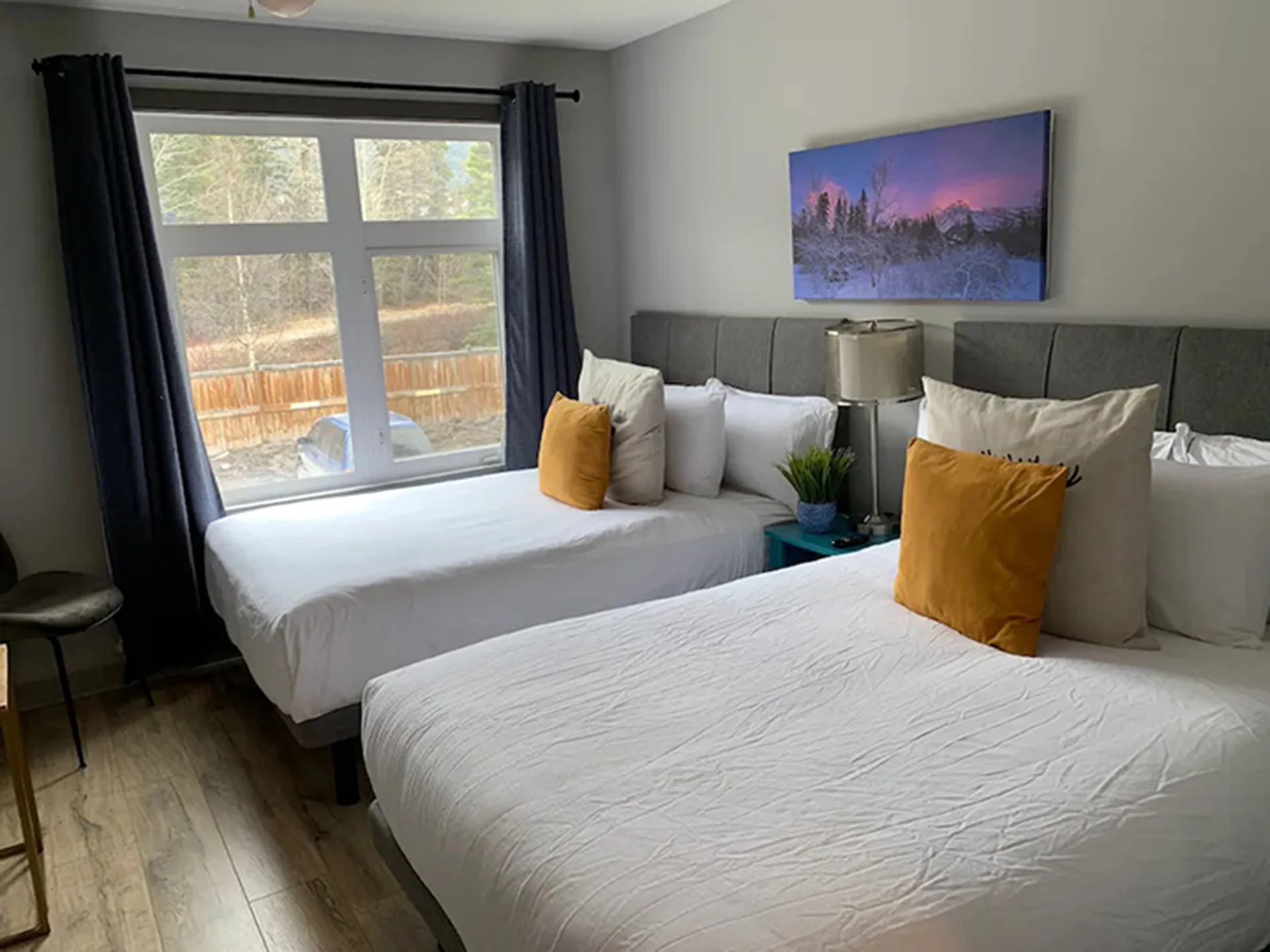 A hotel room with two beds, each with white bedding and mustard-colored pillows. A window with dark blue curtains offers a view of an outdoor area with a wooden fence and trees. A nightstand between the beds holds a lamp and a small potted plant. A painting of a winter landscape hangs above the beds.