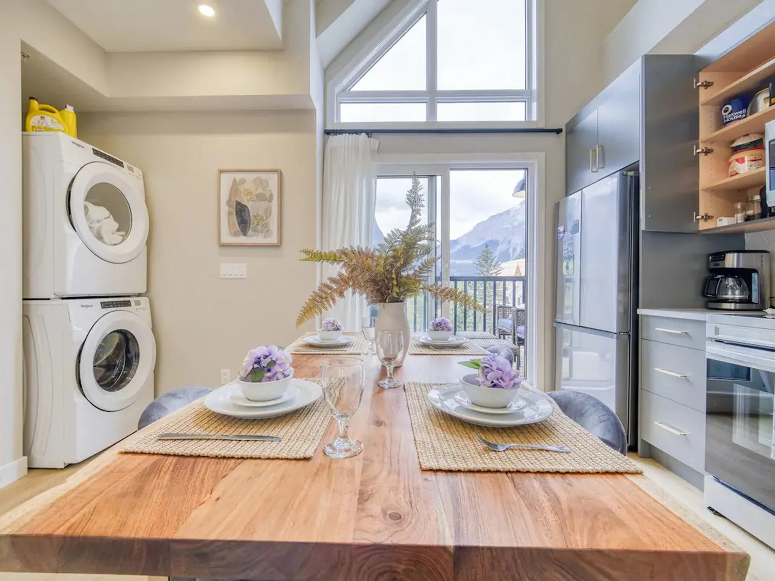 A dining area with a wooden table set for a meal, surrounded by four chairs, with a mountain view through sliding glass doors. The table is decorated with a vase of ferns and purple flowers, and place settings with plates, bowls, wine glasses, and silverware. To the left, there is a stacked washer and dryer, and on the right, kitchen cabinets, a refrigerator, and a coffee maker.