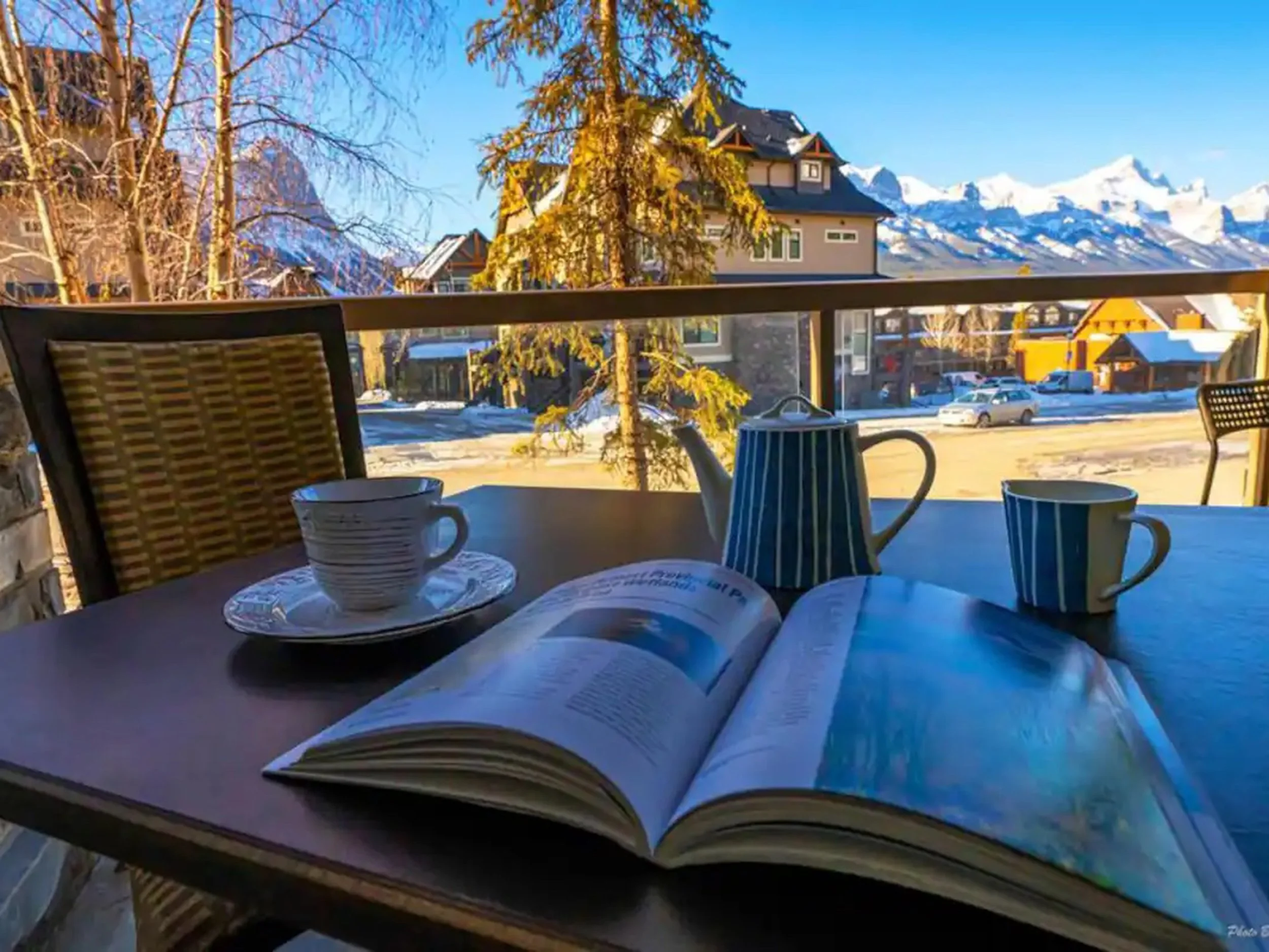 A table on a balcony with a magazine, a striped teapot, two striped mugs, and a cup with a saucer, overlooking a snowy mountain village with houses, trees, and snow-capped peaks in the background.