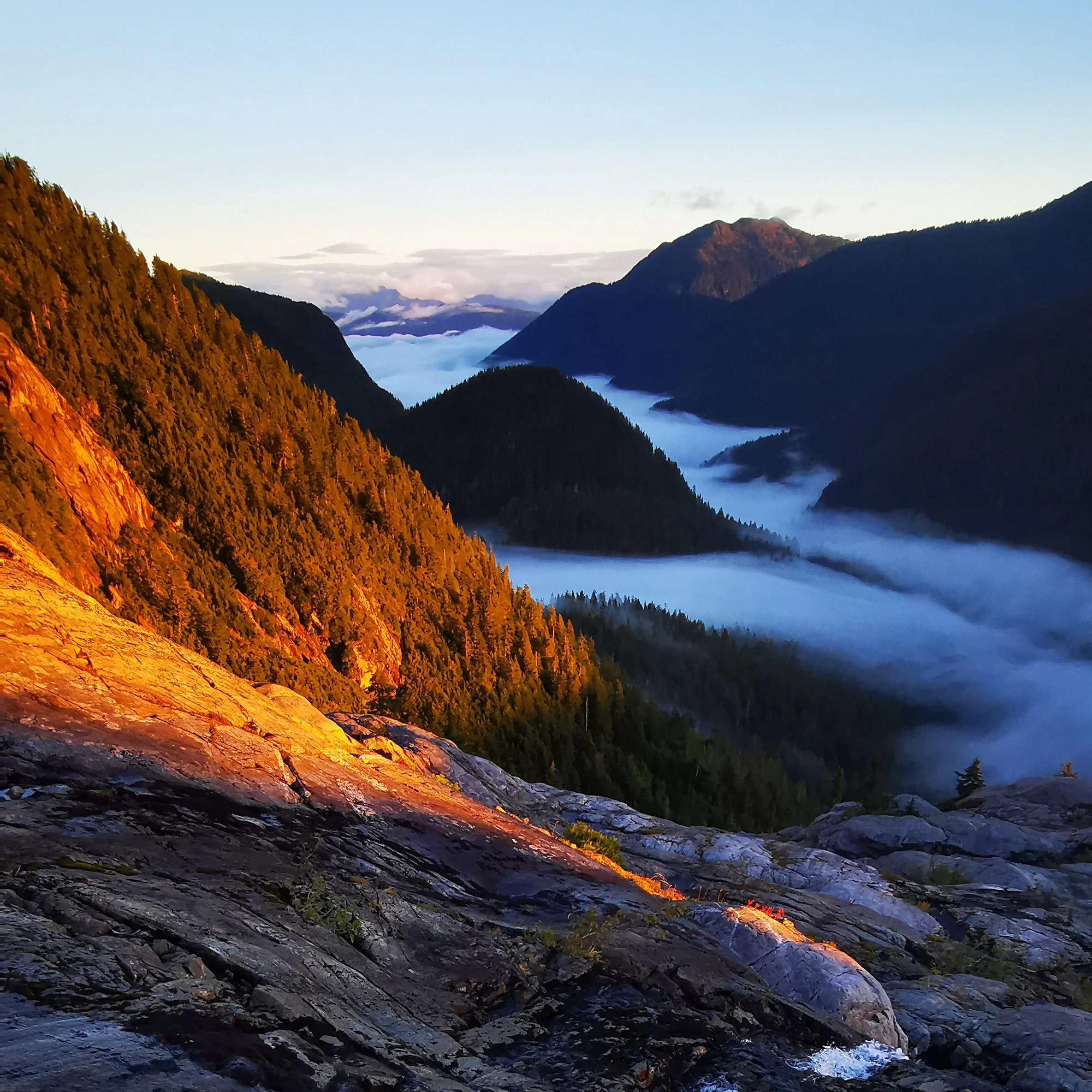 Sunrise on Triple Peak on Vancouver Island, with clouds and fog drifting below the summit and early light illuminating the surrounding mountains.