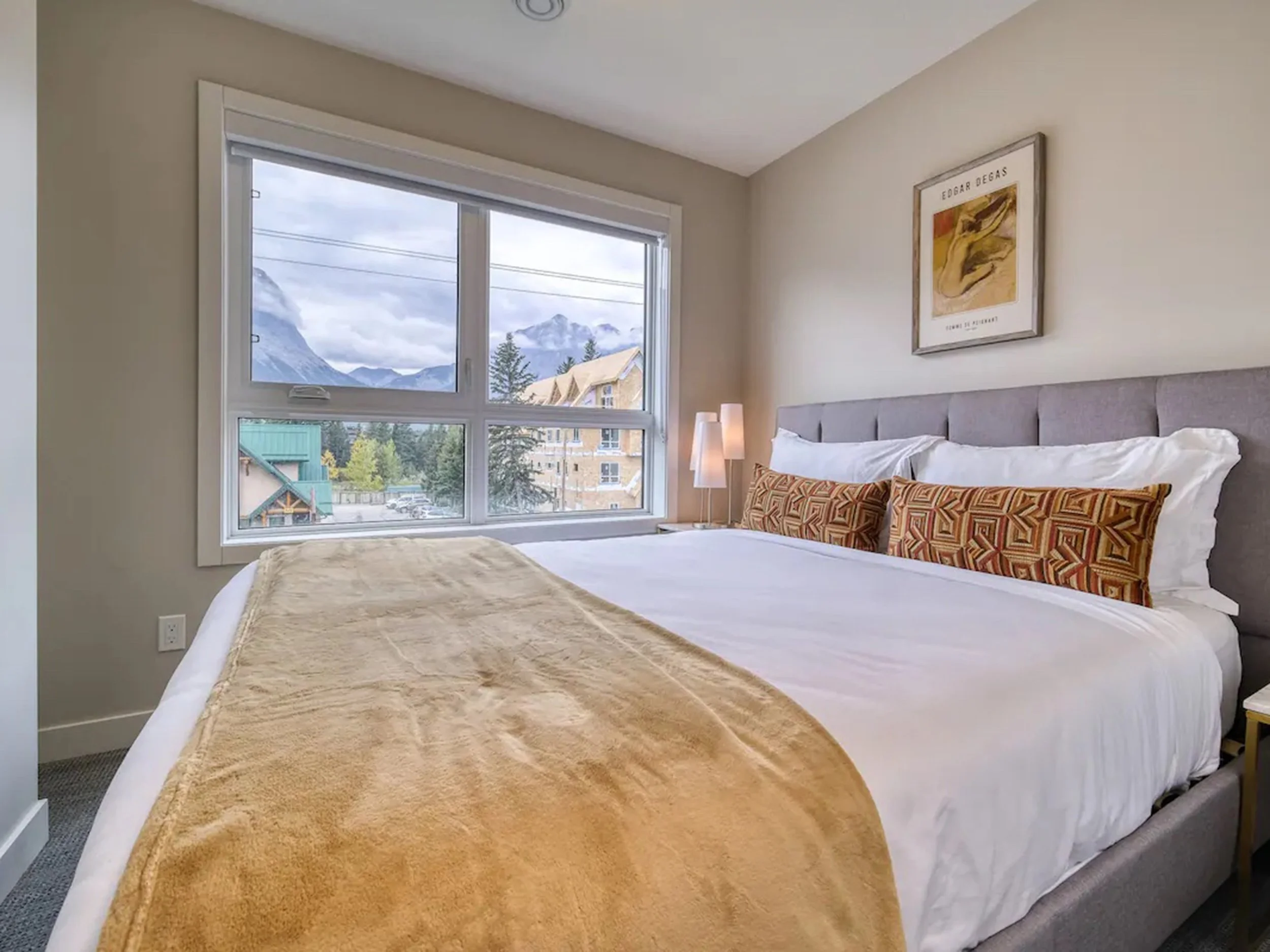 Comfortable bedroom with a large bed, gray upholstered headboard, gold blanket, two patterned pillows, white sheets, side table with modern lamps, large window showing mountains and trees outside, framed artwork on the wall.