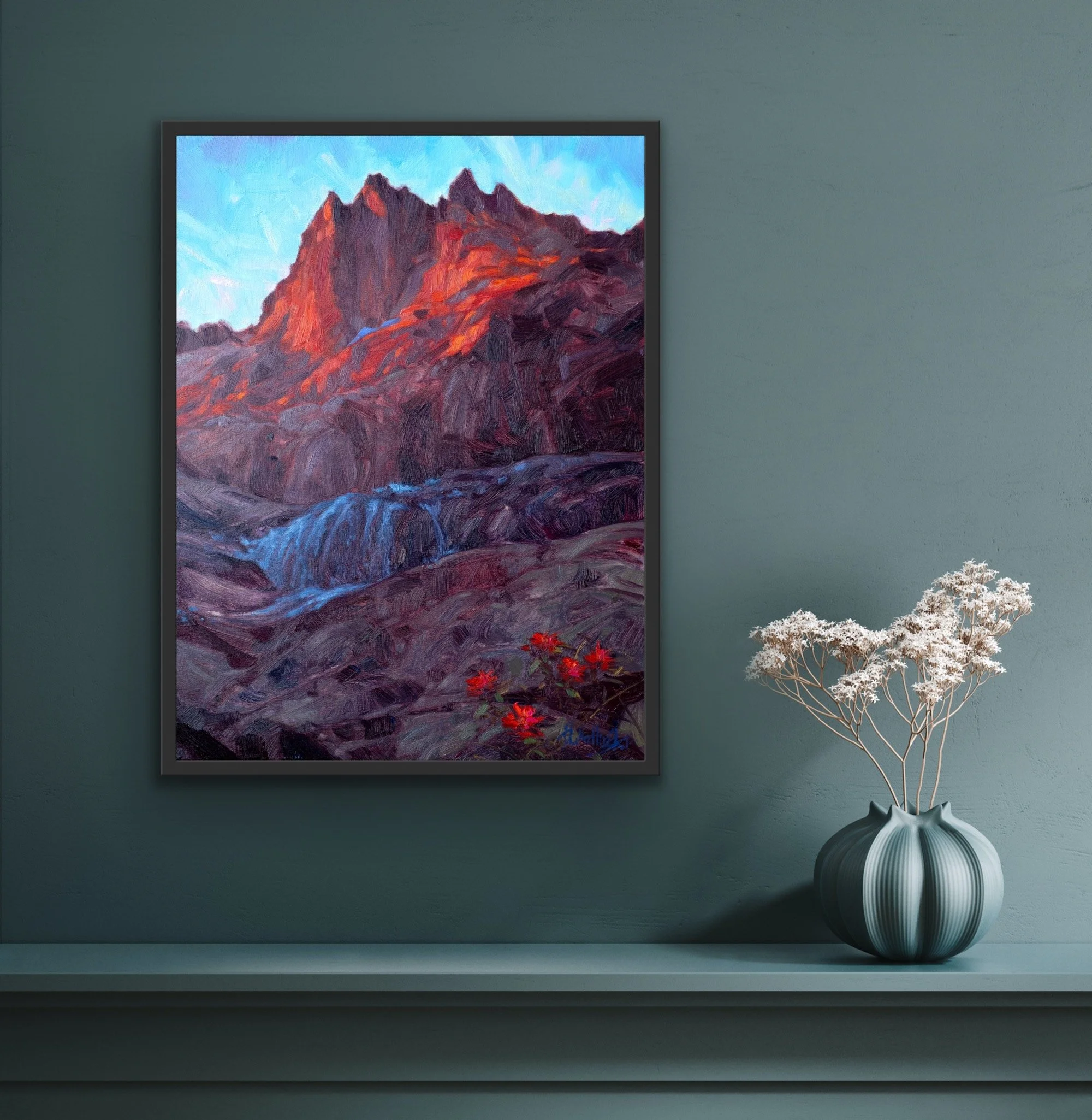 A framed painting of a mountain landscape with red and purple hues, a waterfall, and red flowers, hung on a green wall. A white flowering plant in a black and gray vase is placed on a shelf below the painting.