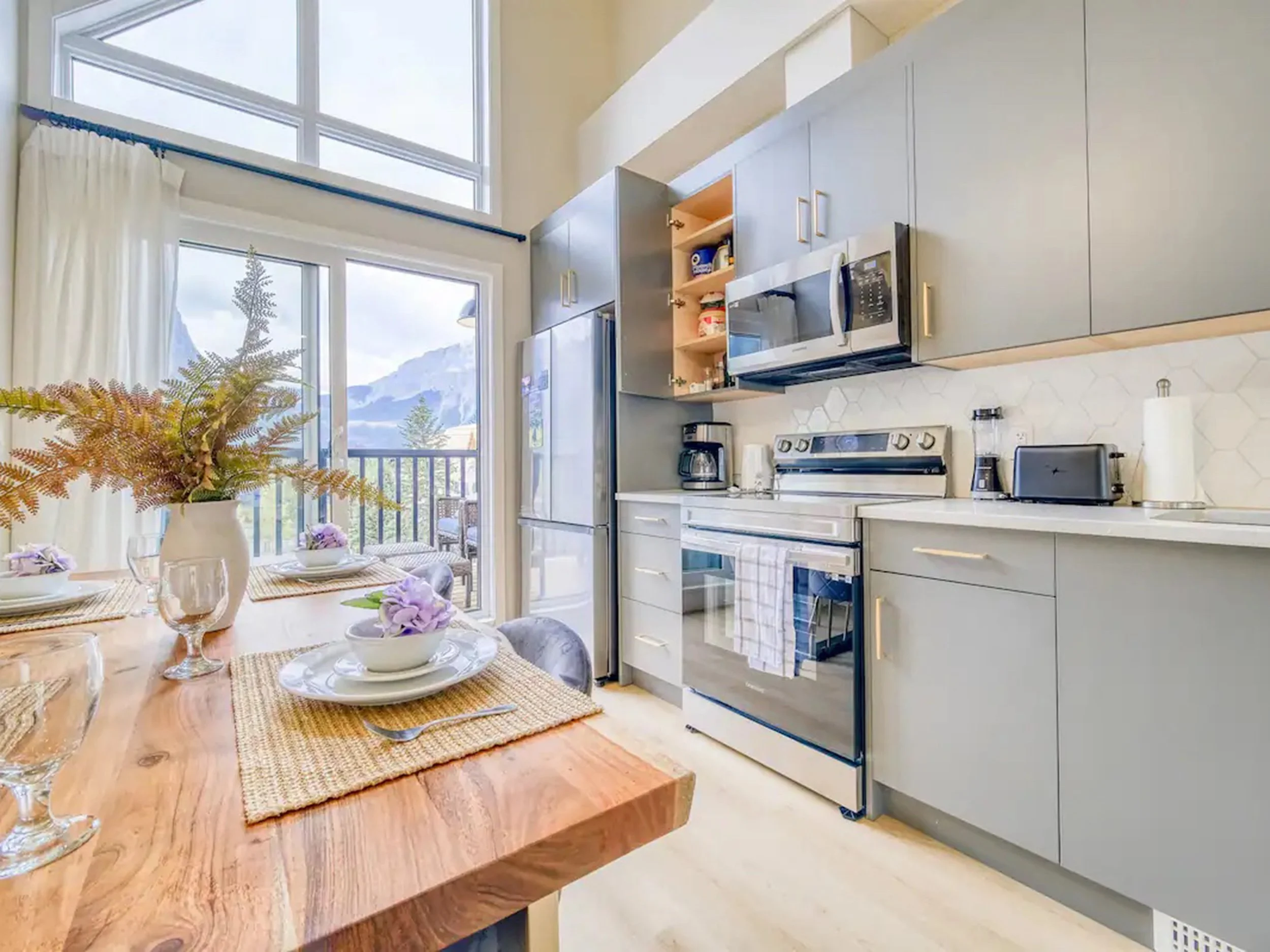 A bright kitchen with light gray cabinets, a stainless steel stove, microwave, and refrigerator. There is a wooden dining table set with plates, bowls, wine glasses, and flowers, near large windows that overlook mountains and a balcony.