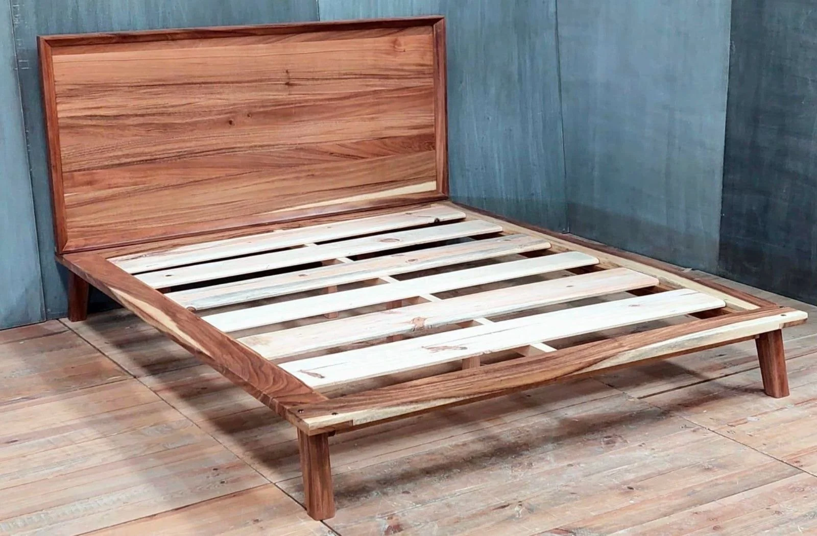 Cama de madera sin colchón, con marco, patas cortas, laterales y cabecera de madera lisa, en una habitación con piso de madera y paredes de madera y tablones.