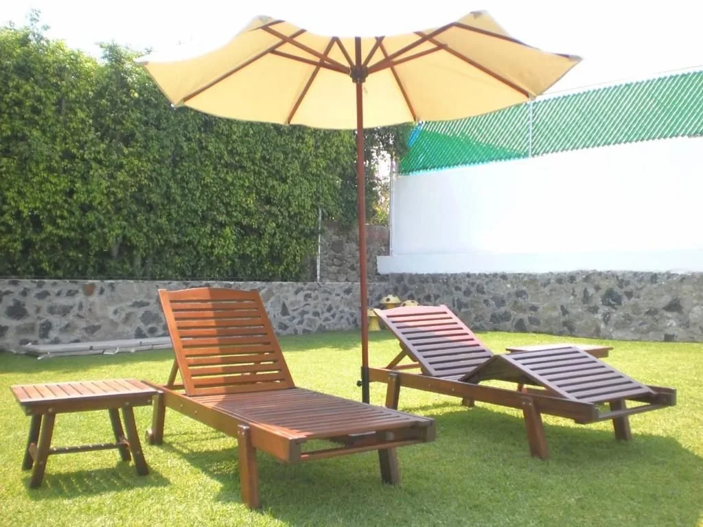 Dos tumbonas de madera bajo una sombrilla en un jardín con césped verde, una cerca de piedra y arbustos.
