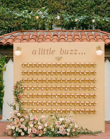 A guest message board titled 'a little buzz...' with rows of wooden clothespins for messages, decorated with pink and white flowers at the bottom and string lights overhead.