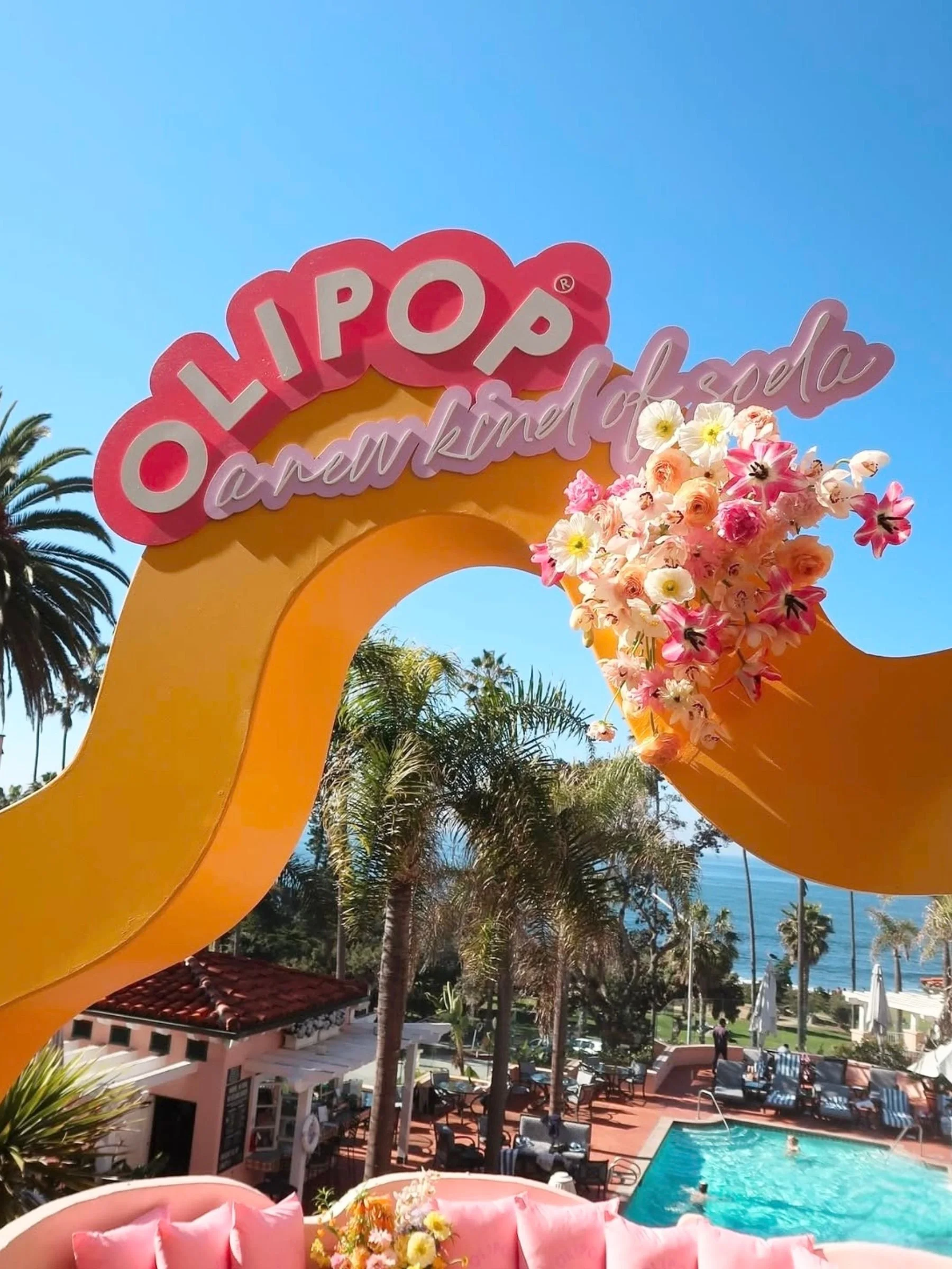 A decorative entrance arch with pink 'Ollipop' sign and cursive text 'amusement of ela' decorated with pink, white, and peach flowers, with a swimming pool, lounge chairs, and palm trees in the background under a clear blue sky.