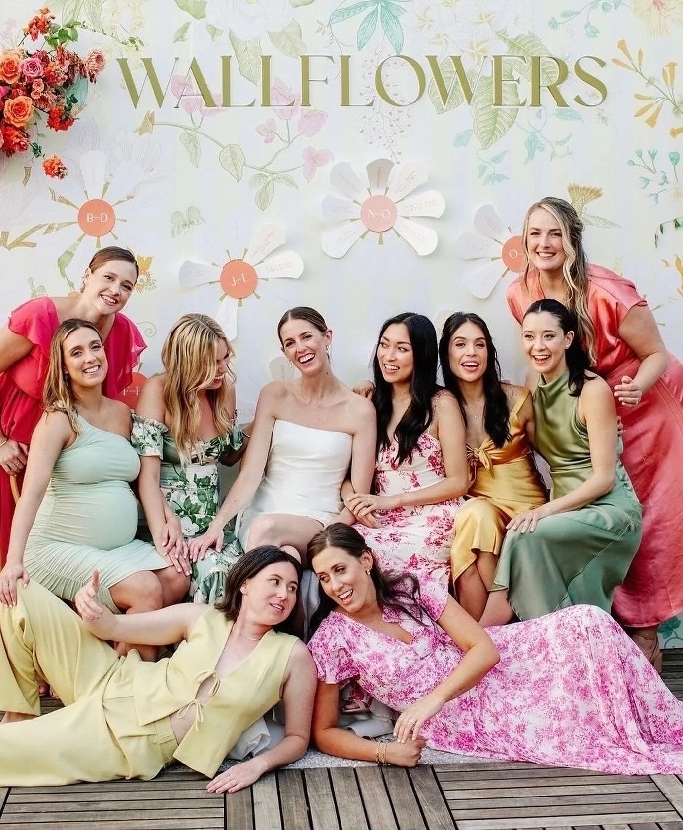 A group of women in colorful dresses sitting and lying on a wooden deck, smiling and posing for a photo, with a floral and greenery backdrop that has the word 'WALLFLOWERS' at the top.