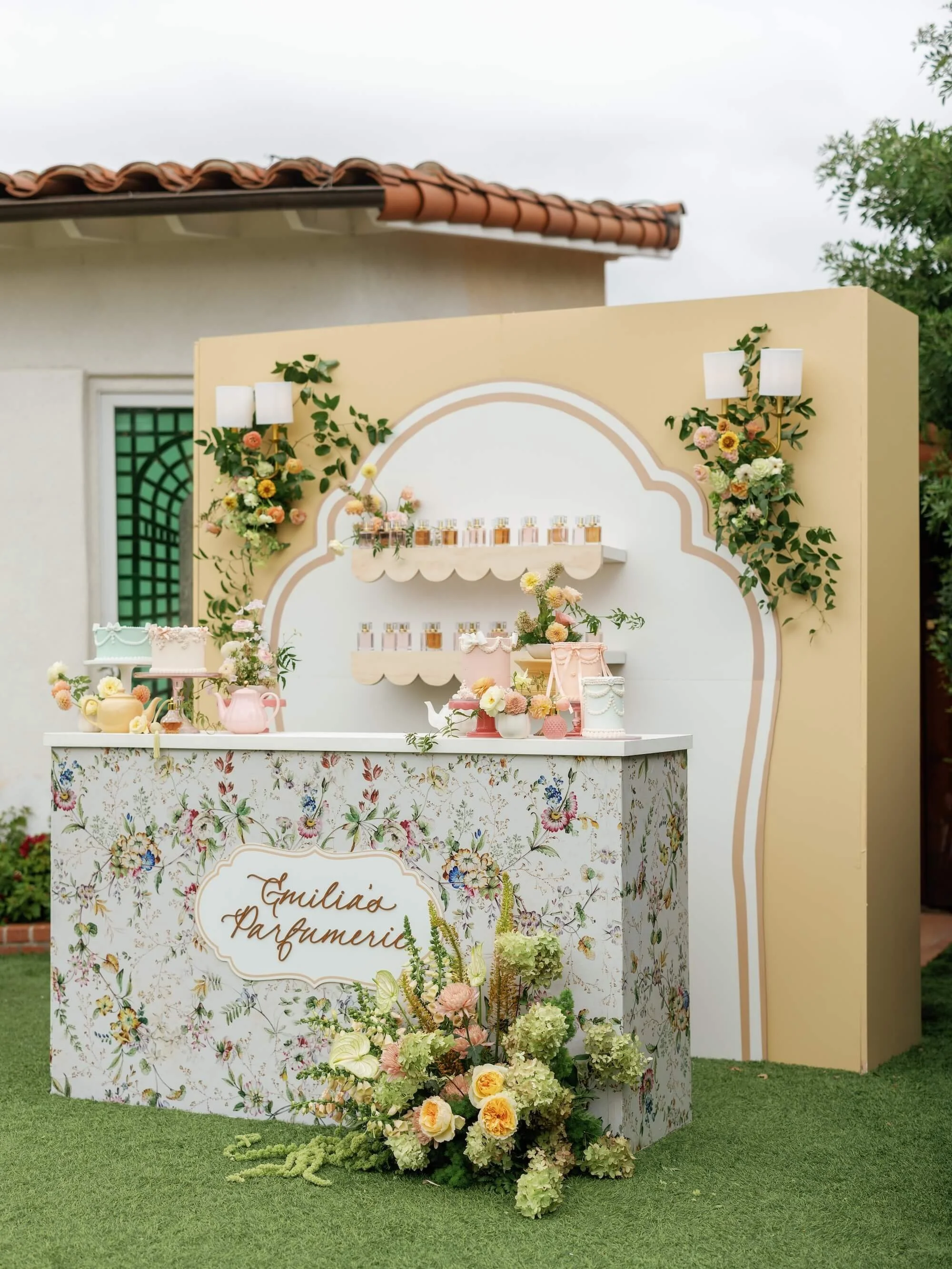 A floral perfume display stand at an outdoor event, decorated with flowers and greenery, marked with the name 'Emilia Parfumerie'.