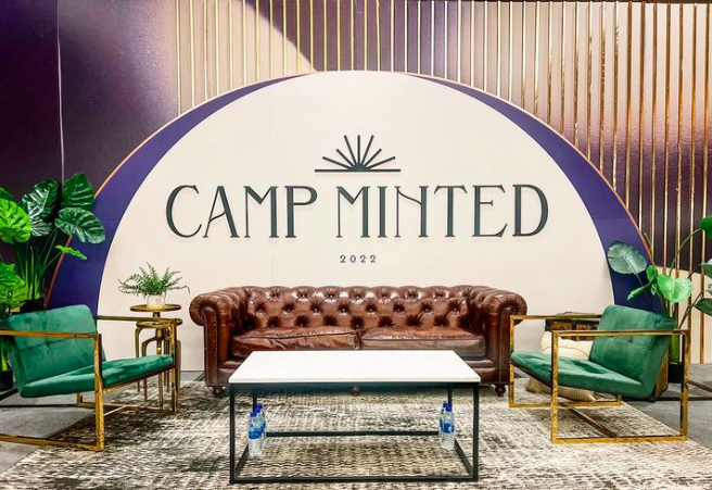 A cozy seating area with a brown leather tufted sofa, two green armchairs with gold frames, a white coffee table, and potted plants, set in front of a large, semi-circular sign reading 'CAMP MINTED 2022' with a stylized sunburst above the text.