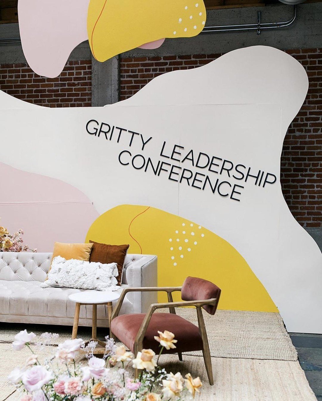Interior of a conference space with a large decorative wall reading "Gritty Leadership Conference," a white tufted sofa with yellow, brown, and white pillows, a wooden armchair with pink upholstery, a small round table, and a backdrop of exposed brick wall and industrial piping, with pink and white flowers in the foreground.