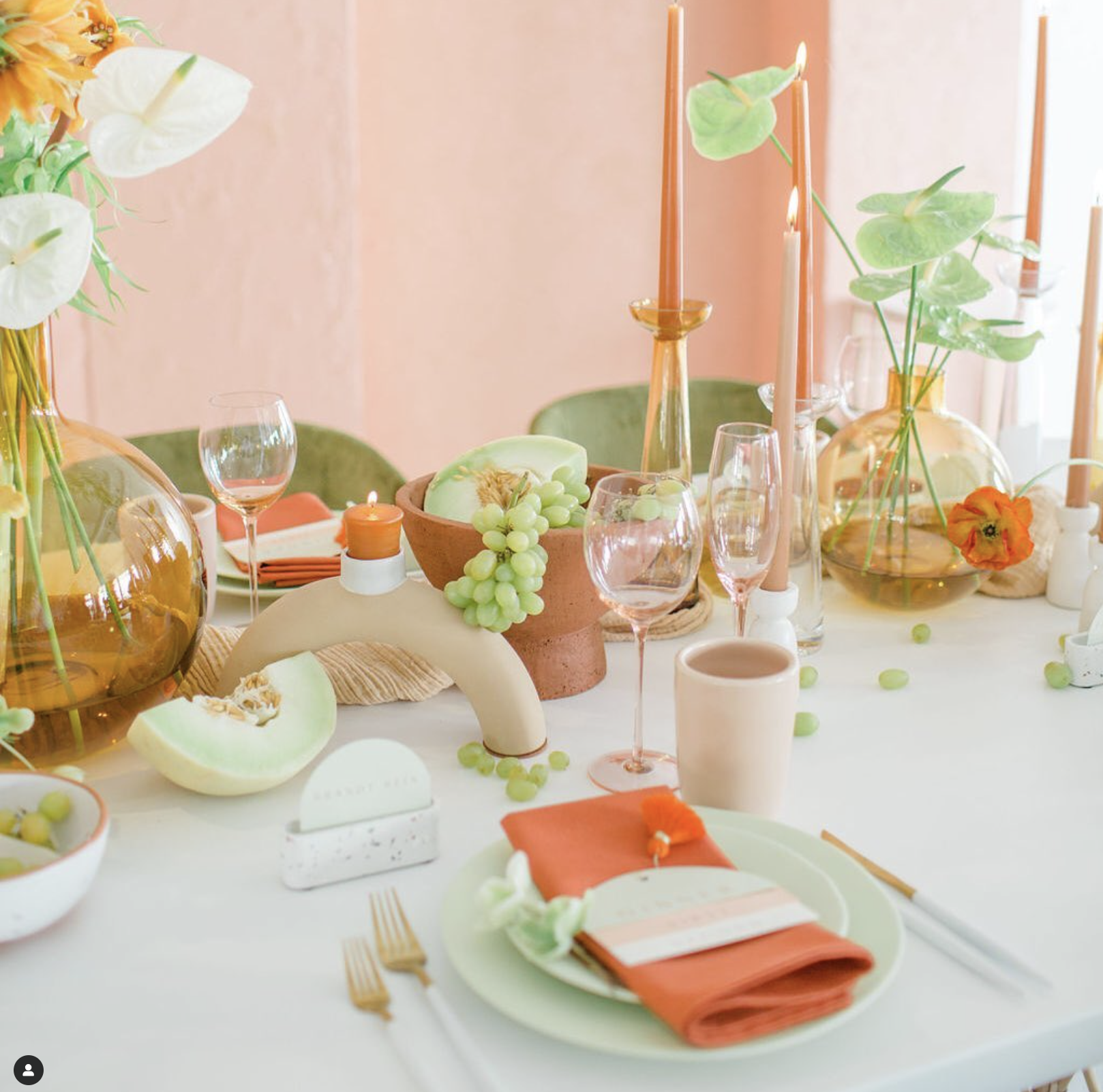 A tablescape with pastel-colored dishes, candles, flowers in vases, green grapes, cut melons, and glassware, arranged on a white table with a pink background.