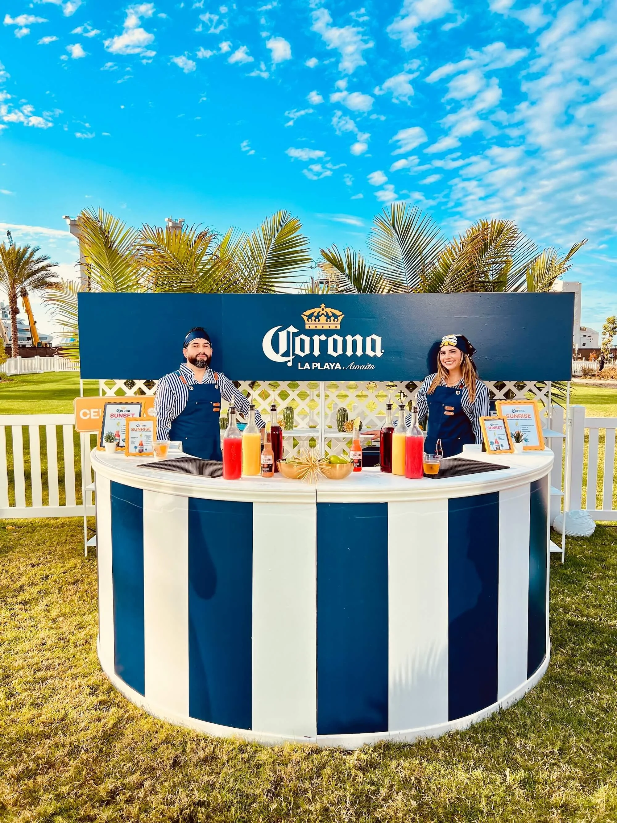 A promotional stand with a blue and white striped design, featuring two staff members in navy aprons and headbands. The stand displays bottles with colorful drinks and promotional signs for Corona Sunset and Corona Sunrise, set outdoors with palm trees and clear blue sky in the background.