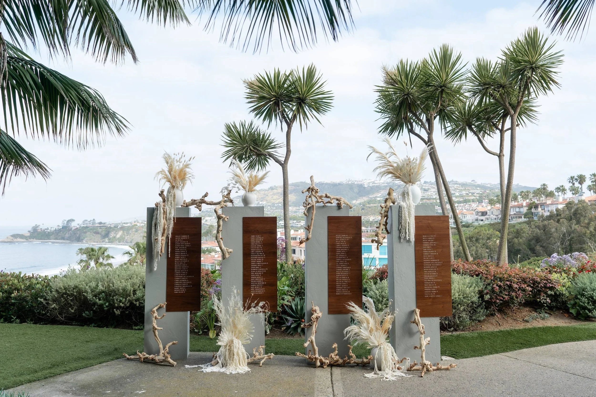 Memorial monument with wooden plaques, decorated with driftwood and feathery plants, overlooking the ocean with palm trees and hillside houses in the background.