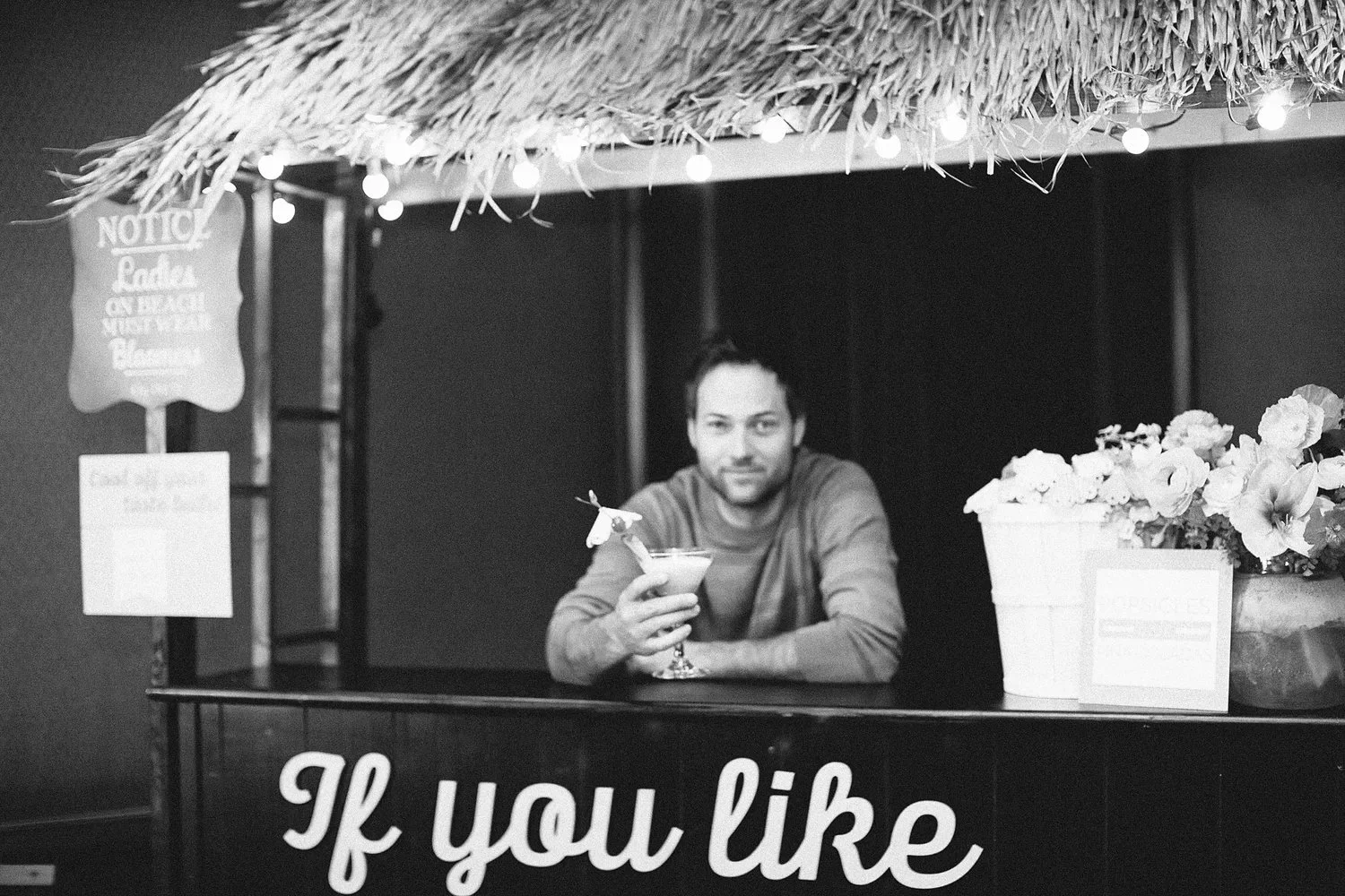 A man behind a small stand with a thatched roof, holding a cocktail with a small umbrella, in black and white. There are light bulbs hanging from the roof and a flower arrangement on the right side. The front of the stand has partially visible text that reads, 'If you like'.