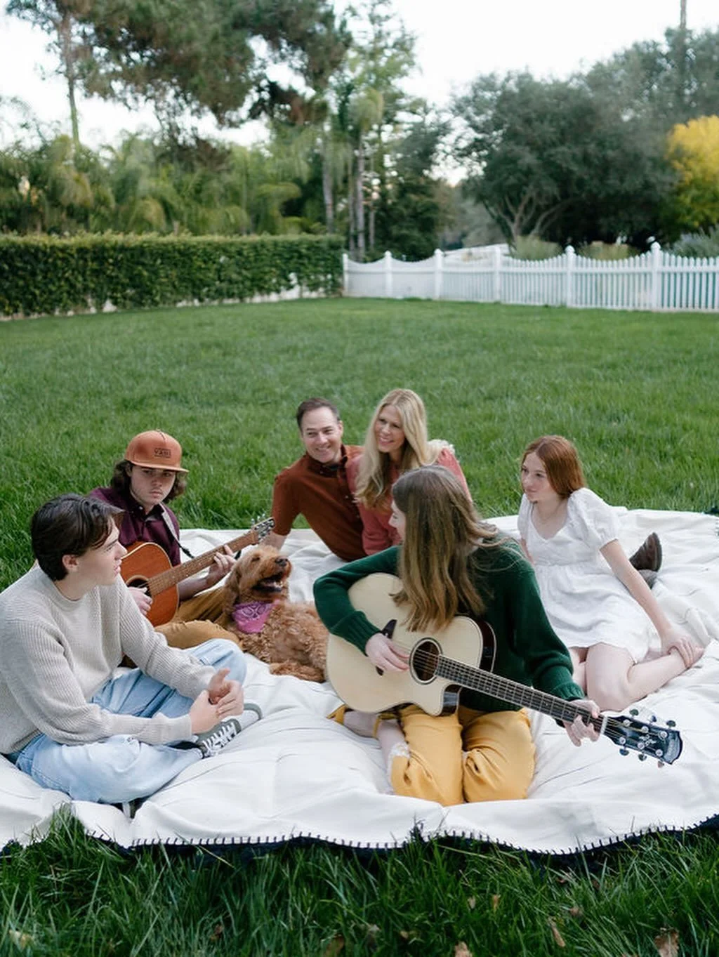 A family sitting on a white blanket on the grass, playing guitars, and enjoying a picnic in a backyard with trees and a white fence.